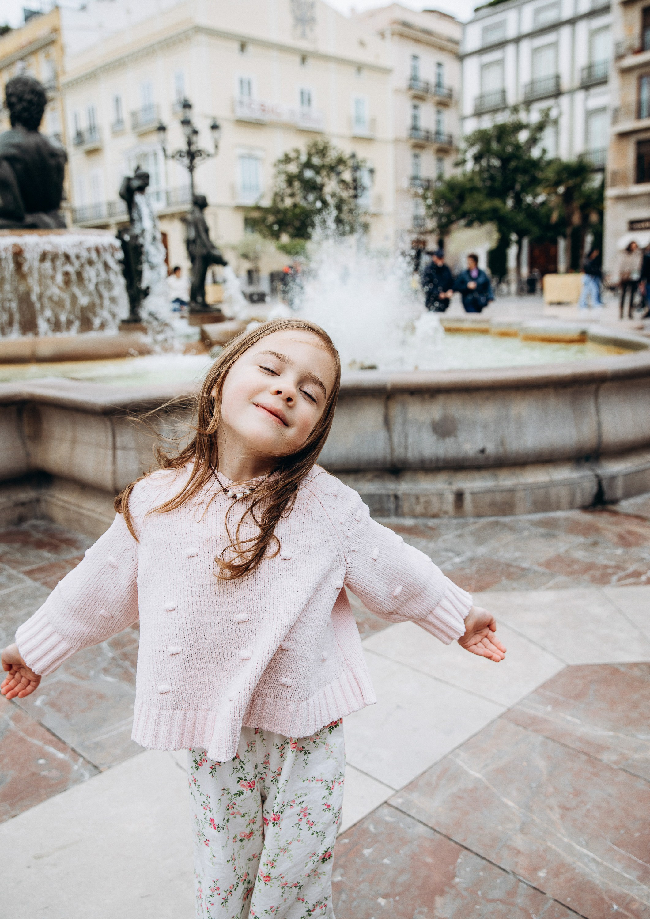 Семейная фотосессия в Валенсии, Испания, где маленькая девочка радостно кружится возле исторического фонтана в очаровательной городской локации, снято при естественном свете для игривого, искреннего и lifestyle-семейного момента — идеально для семейных фотосессий в Валенсии и по всей Испании.