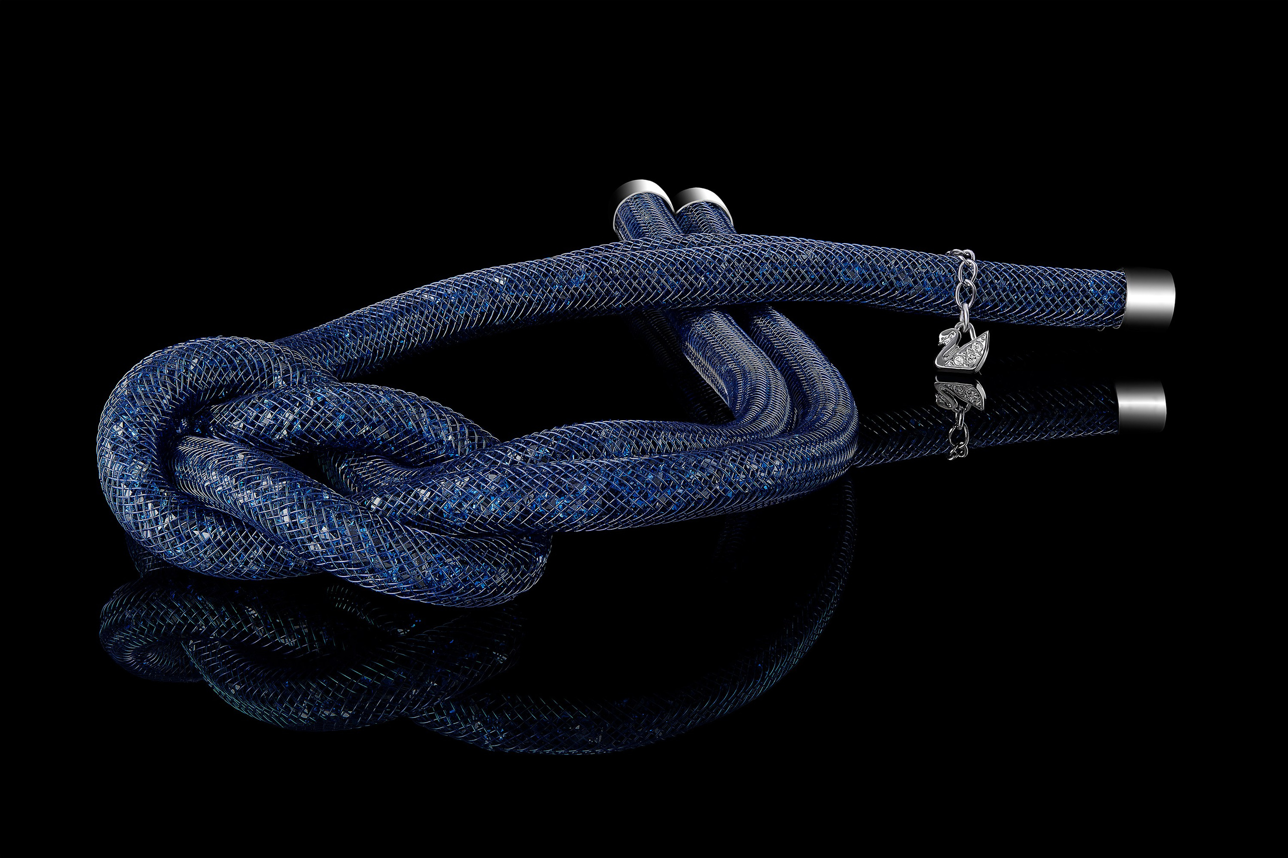 Blue Swarovski Stardust necklace with decorative knot and swan charm on black reflective background, close-up product photography of jewelry.