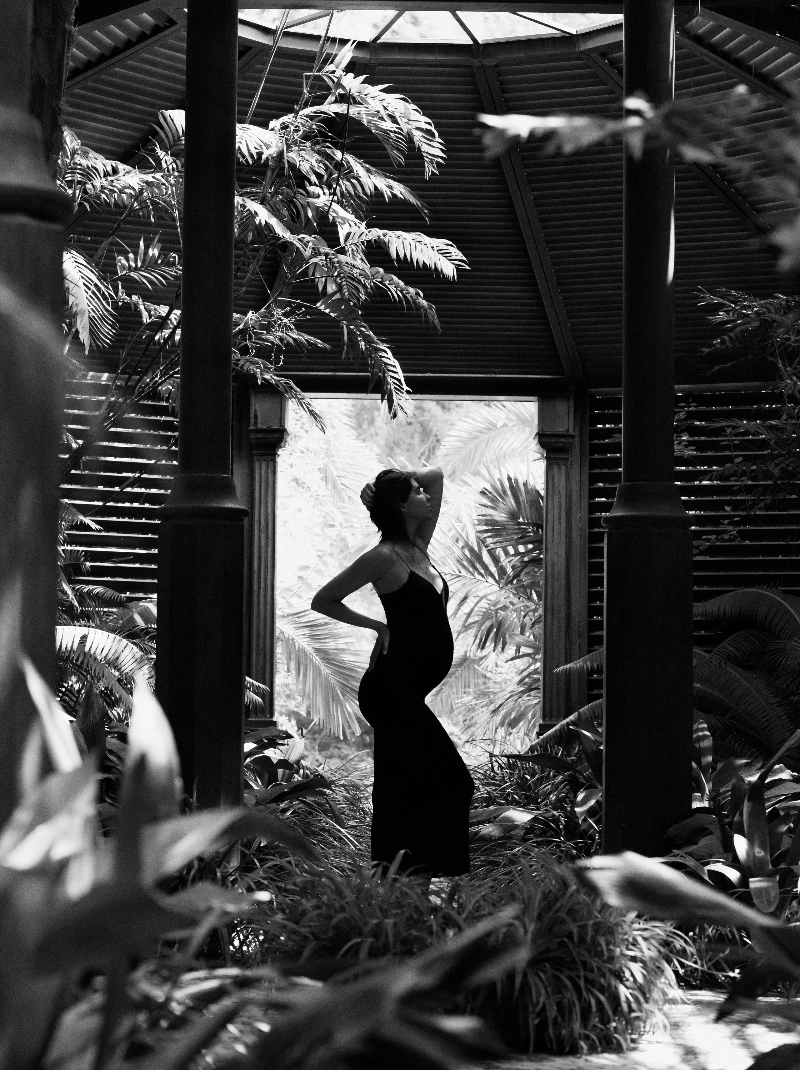 Retrato artístico en blanco y negro de una mujer embarazada posando con gracia dentro de un invernadero tropical en Valencia, España, rodeada de columnas arquitectónicas y luz natural filtrada entre las hojas.