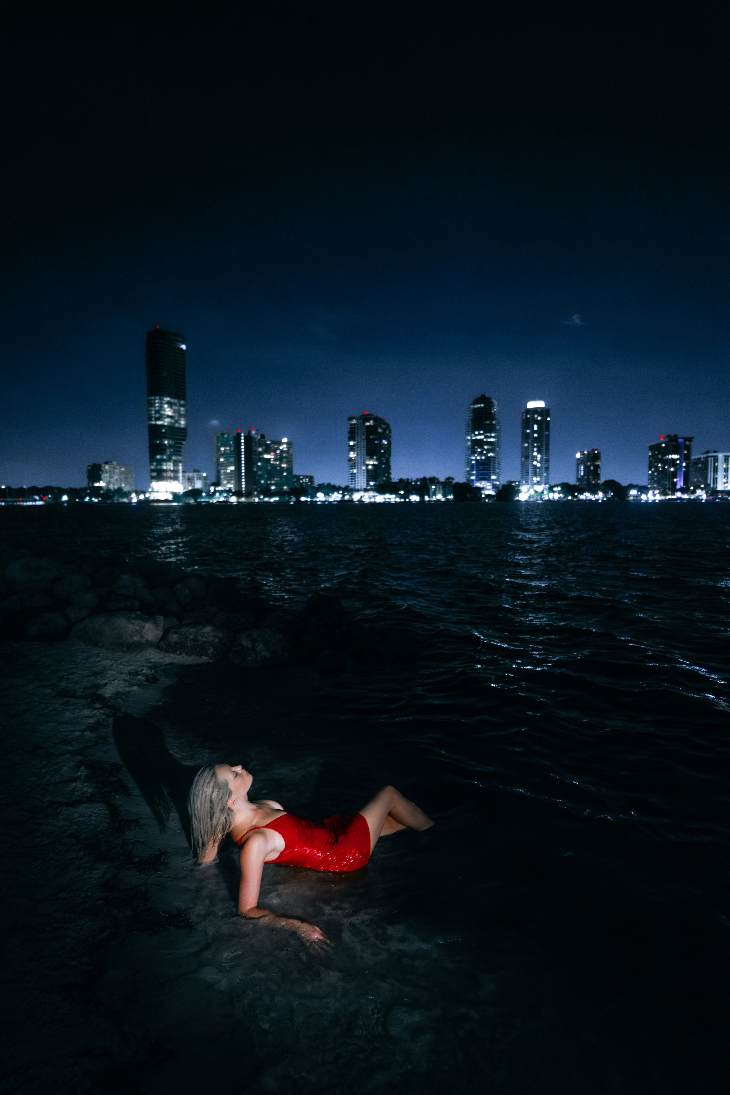 Model sits on shoreline rocks with bare feet in the water, Miami’s night skyline reflecting across the bay. Cinematic control of color and shadow.
