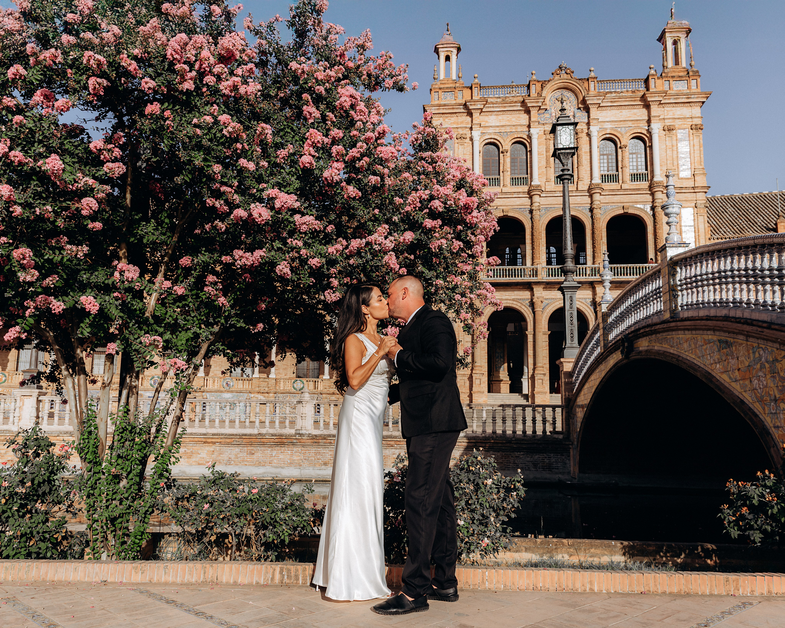 Romantic wedding photo of a couple sharing a kiss under blooming pink flowers at Plaza de España in Sevilla, Spain — a breathtaking location perfect for couples seeking vibrant and unforgettable wedding photoshoots in Seville and southern Spain.