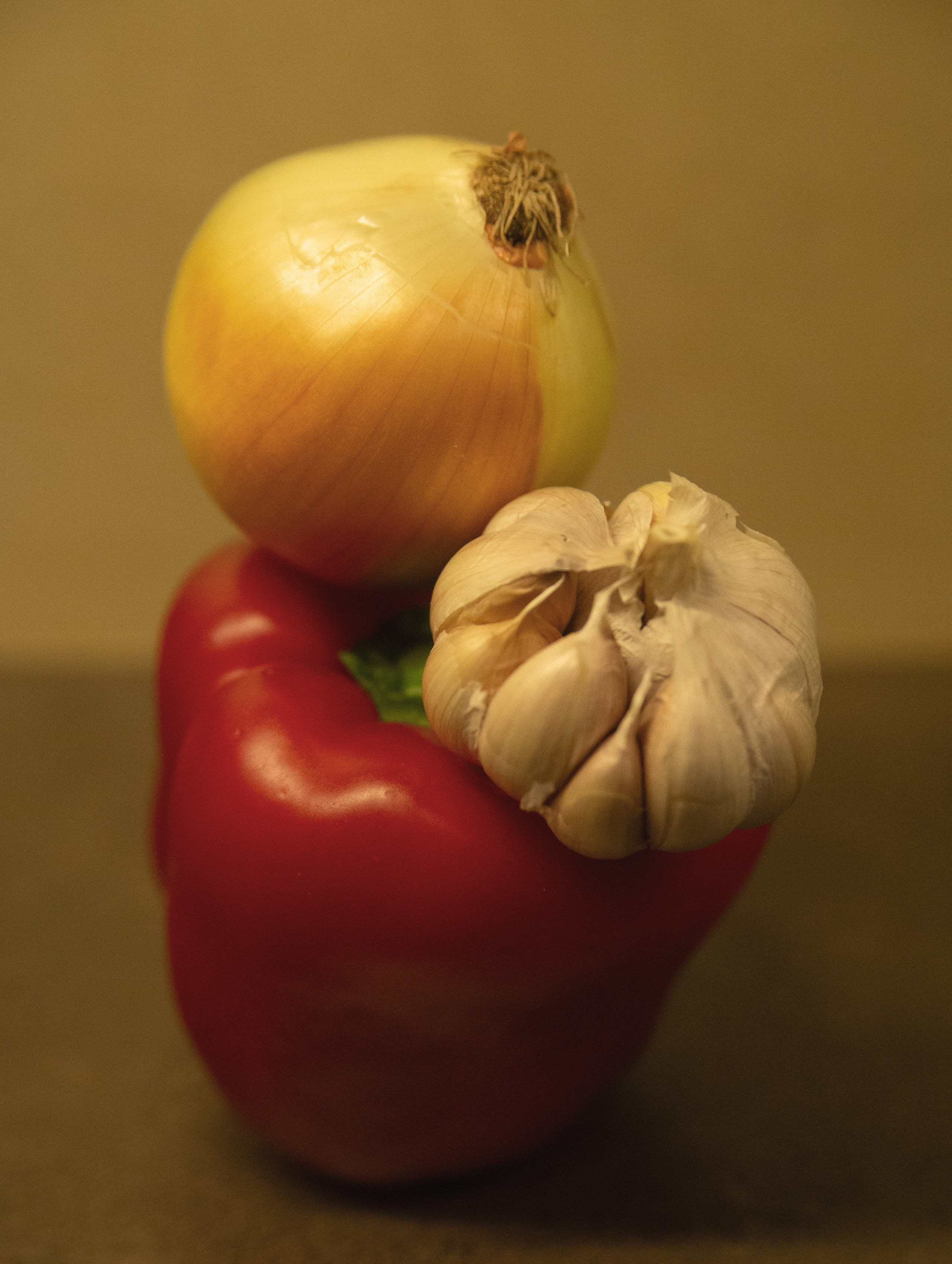 Balanced still life of a red bell pepper, garlic bulb, and yellow onion stacked together, showcasing natural color harmony and rustic simplicity