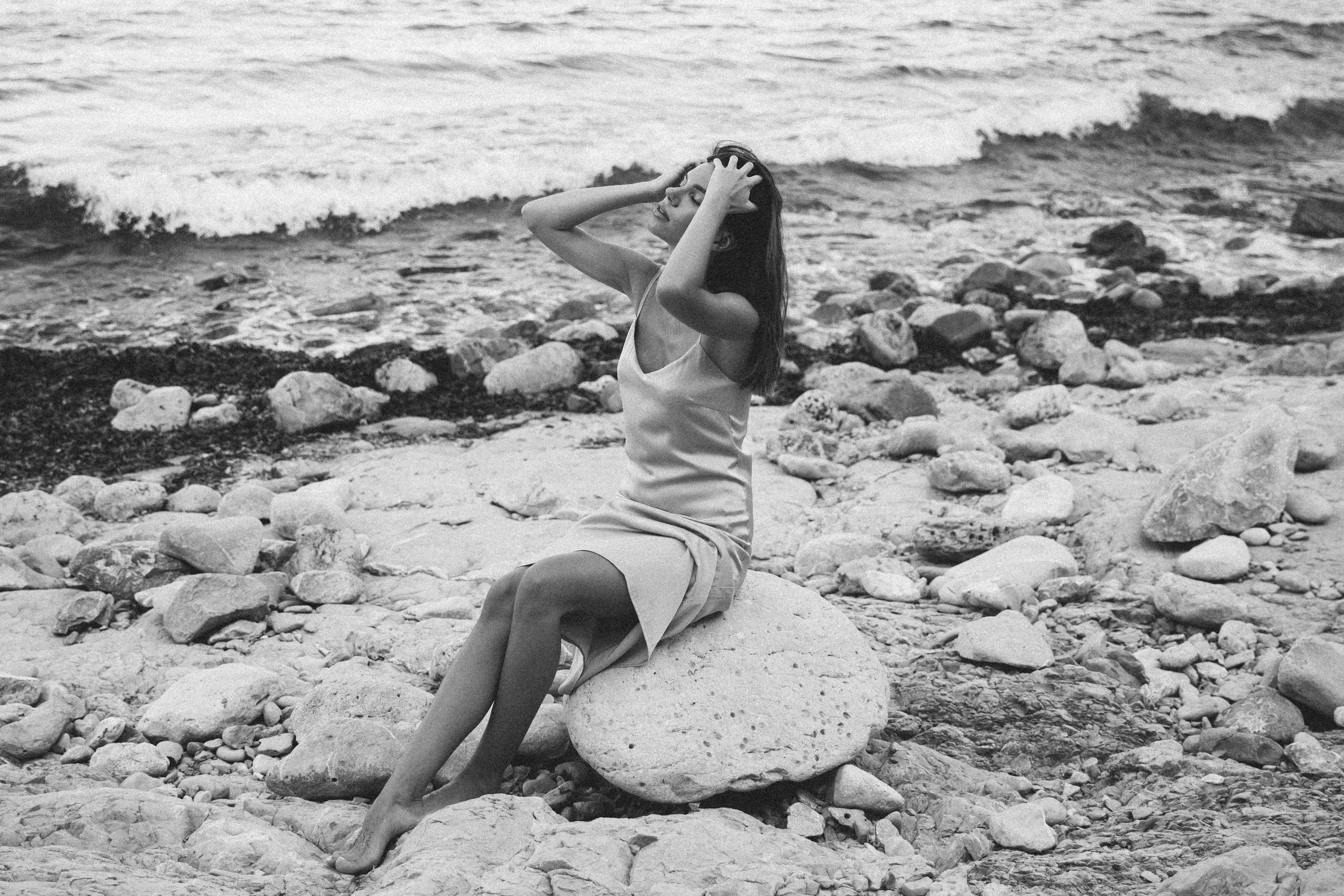 Женская фотосессия в Валенсии, Испания, с художественным чёрно-белым портретом женщины, сидящей босиком на прибрежных камнях и аккуратно придерживающей волосы, на фоне средиземноморских волн, снятым в естественном свете — идеально для пляжных женских фотосессий в Валенсии, эмоциональных прибрежных портретов и профессиональной женской фотографии по всей Испании.