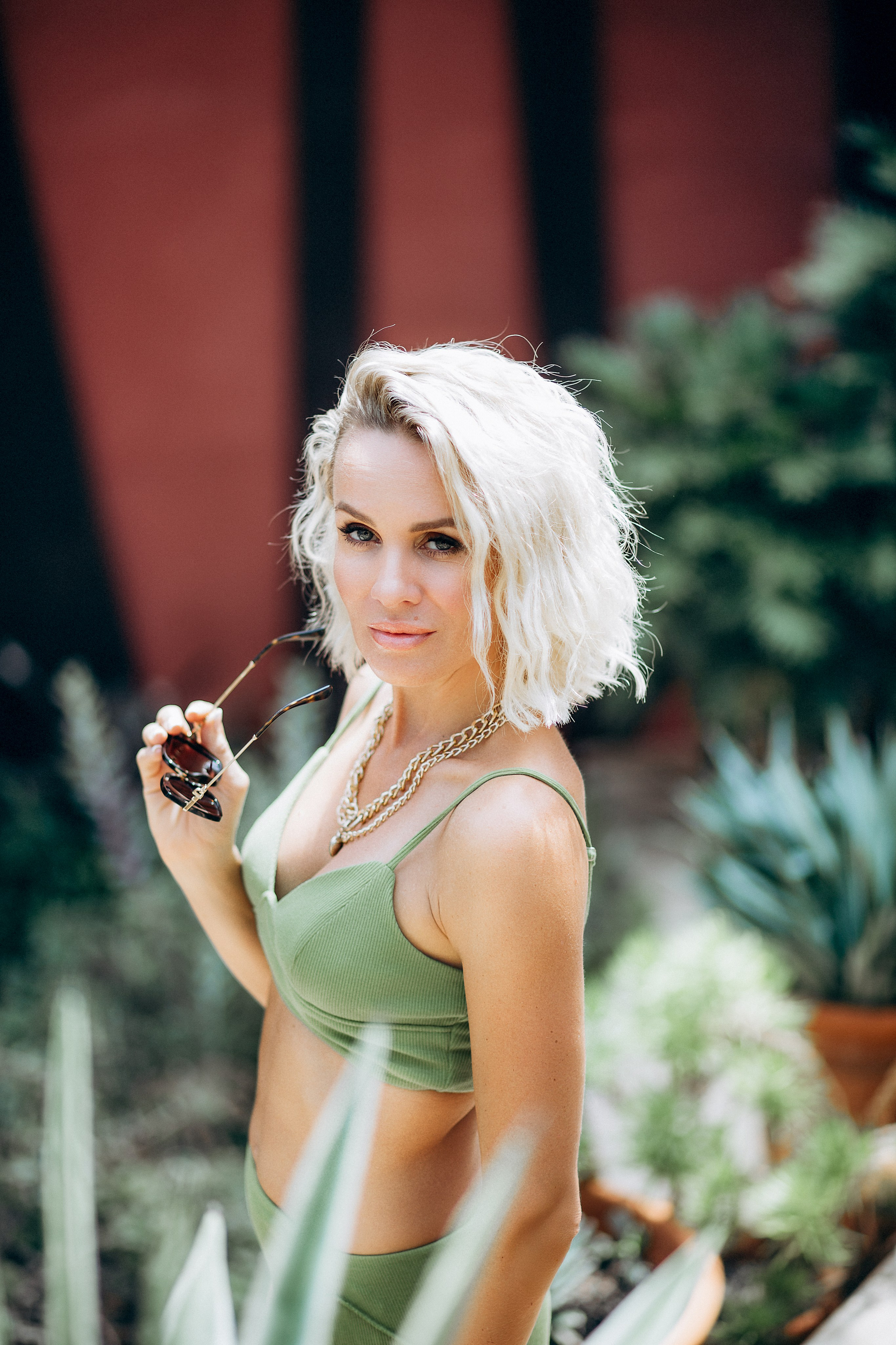 Модная летняя фотосессия в Торревьехе, Испания — блондинка в зелёном костюме с солнцезащитными очками в окружении тропических растений и ярких цветов. Прекрасный вариант для стильной современной портретной съёмки в Торревьехе и регионе Коста-Бланка.