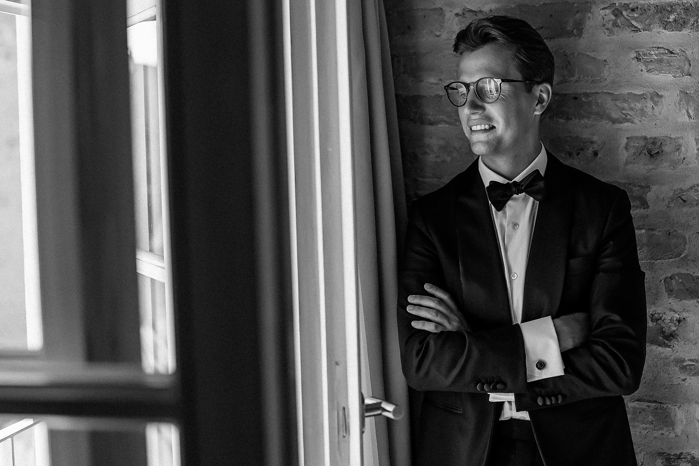 The groom in a tuxedo with a bow tie is standing with arms crossed, leaning against a brick wall near a window with curtains. The image is in black and white.