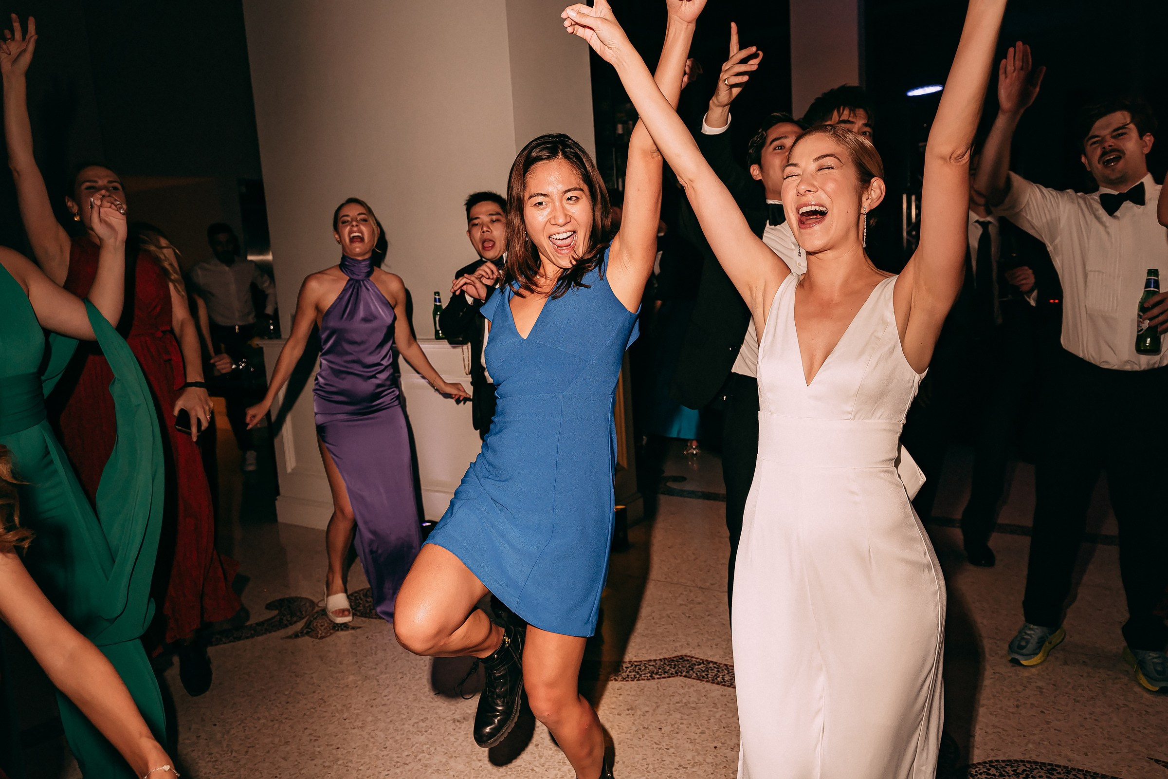 Two women, one in a blue dress and the other in a wedding gown, dancing with arms raised and smiling.