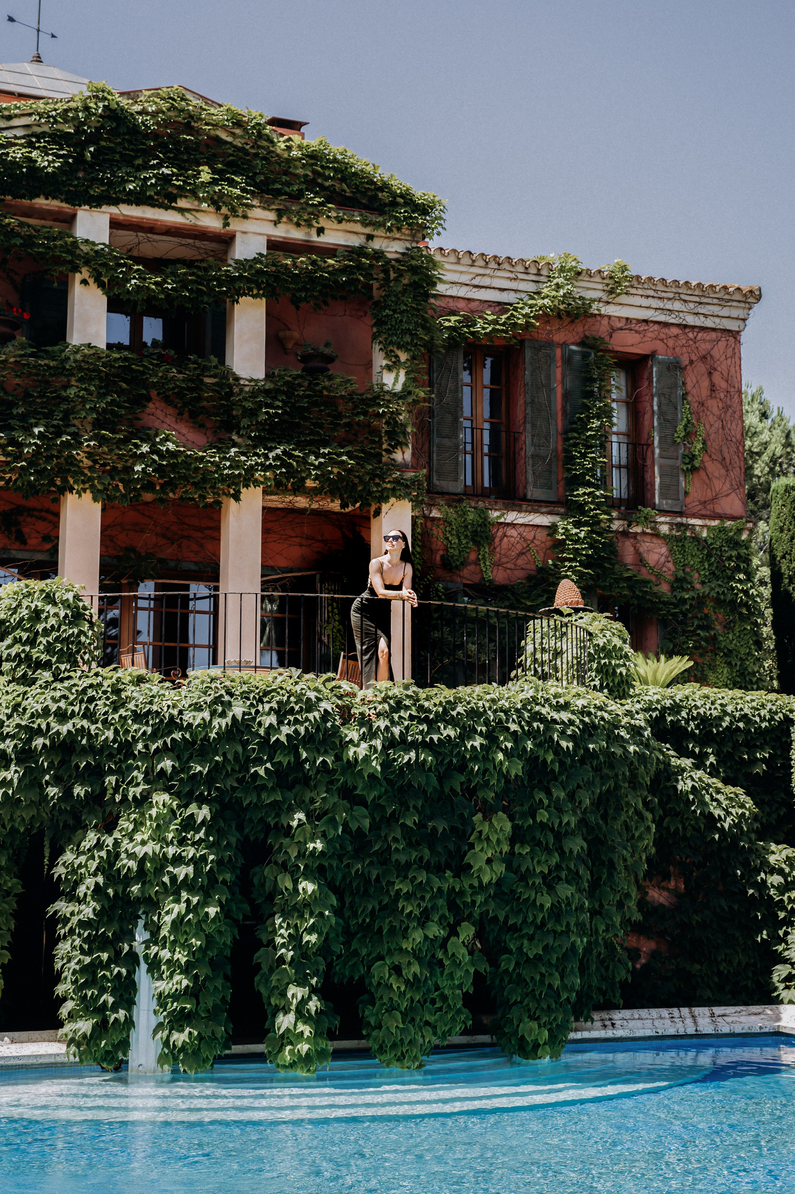 Шикарная женщина в солнцезащитных очках и стильном черном платье позирует на балконе, увитом виноградной лозой, над спокойным бассейном в средиземноморской вилле в Барселоне, Испания — роскошная портретная фотография, сочетающая элегантность, природу и архитектурное очарование, идеально подходит для портретных фотосессий в Барселоне и Испании.
