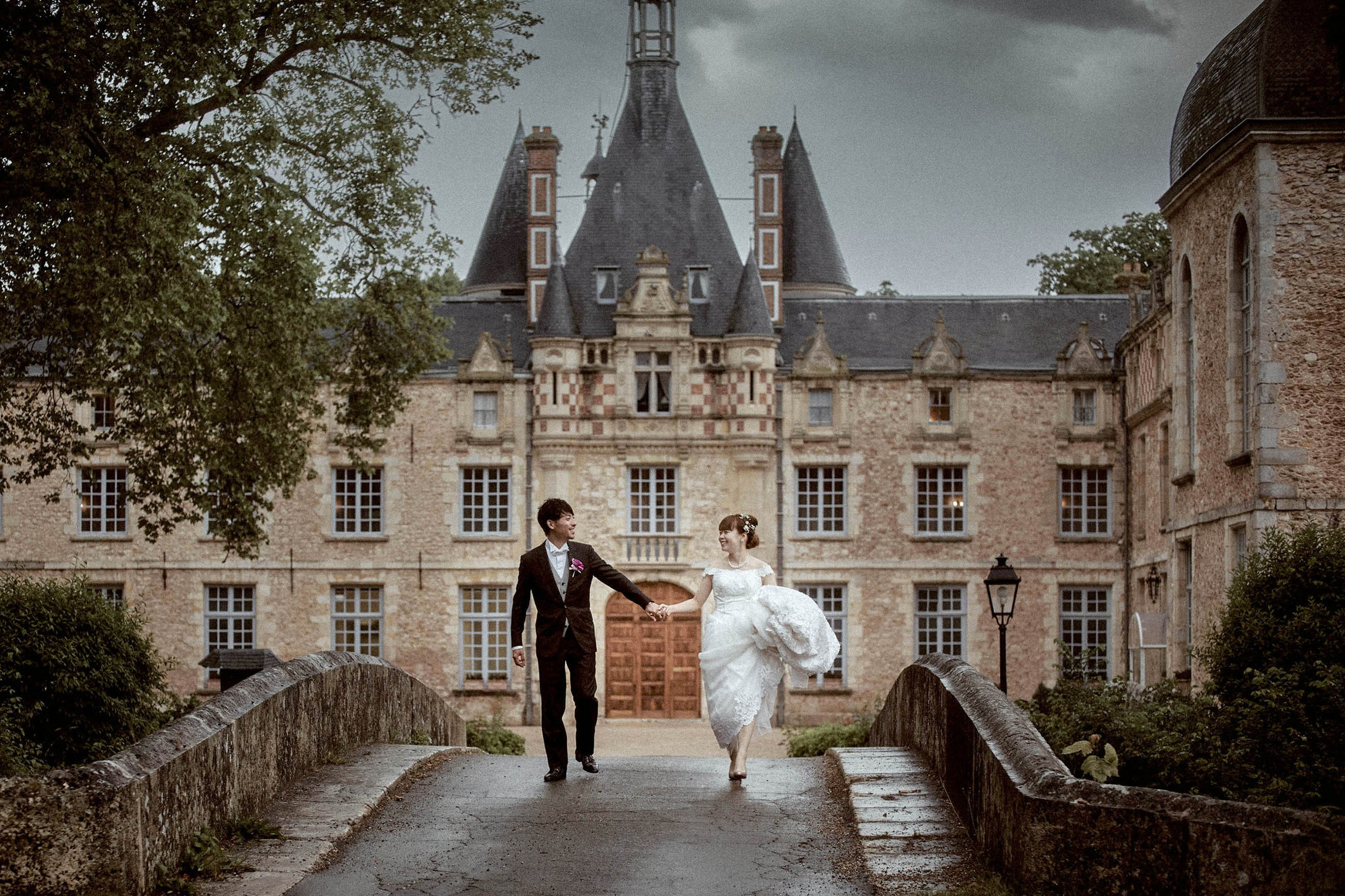 Smiling newlyweds stroll hand in hand in front of the historic Chateau d'Esclimont in France during their wedding day.