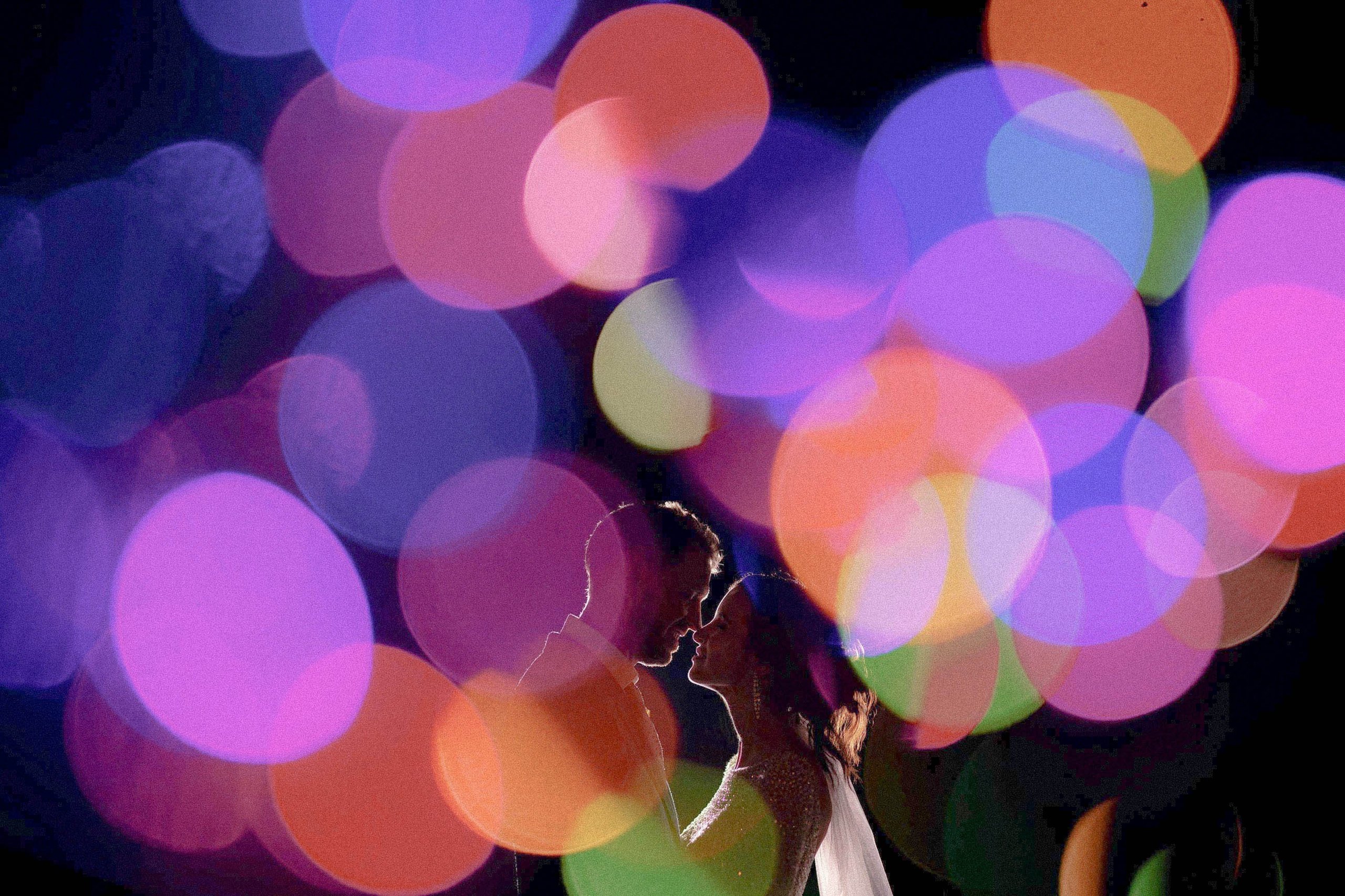 A smiling bride & groom are silhouetted against a colorful foreground of bokeh balls in various colors.