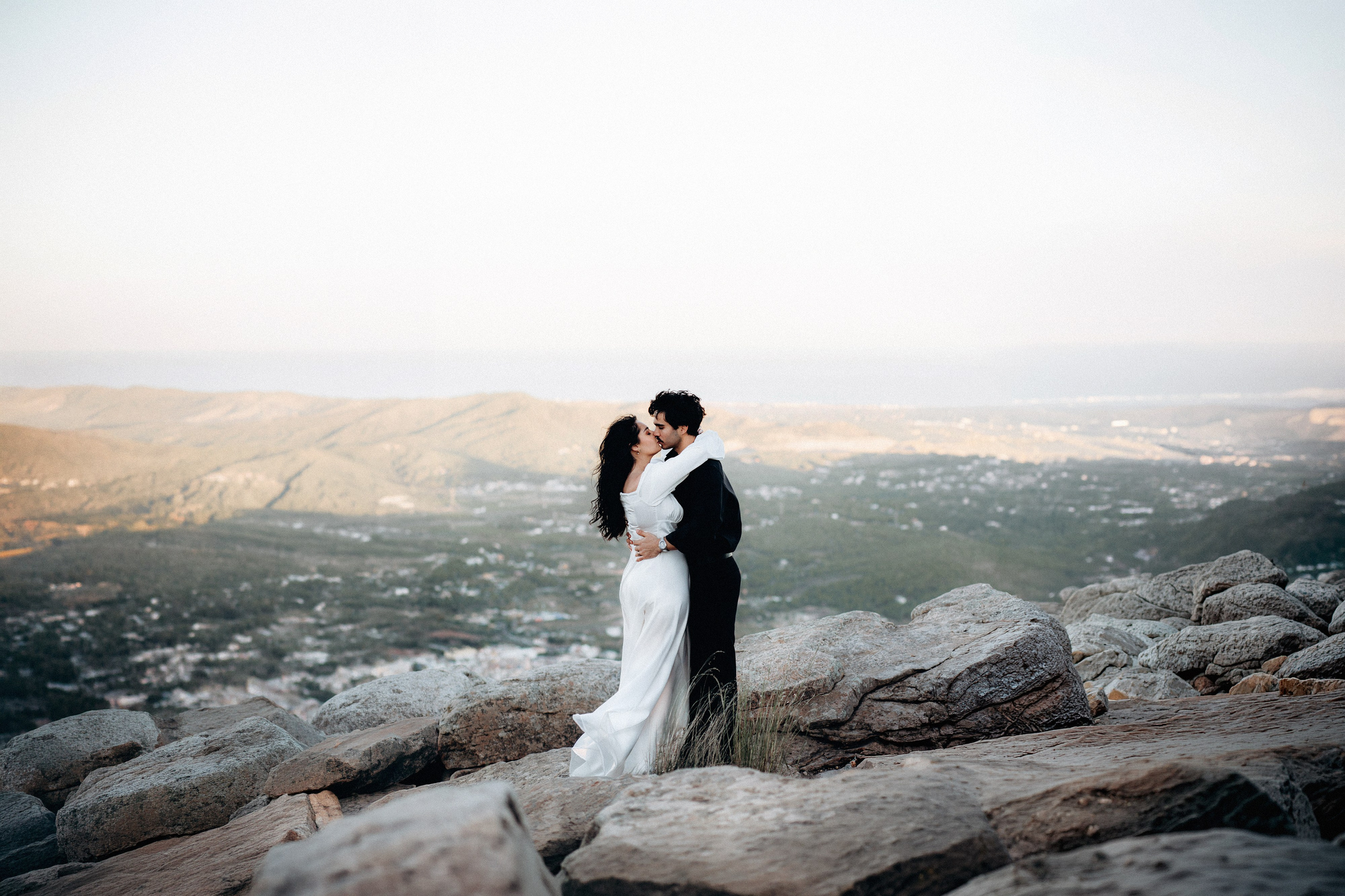 Bride and groom standing on a rocky mountain viewpoint during their intimate elopement in Barcelona, Spain, overlooking a scenic valley at sunset. The destination wedding portrait highlights the dramatic landscape and romantic atmosphere.