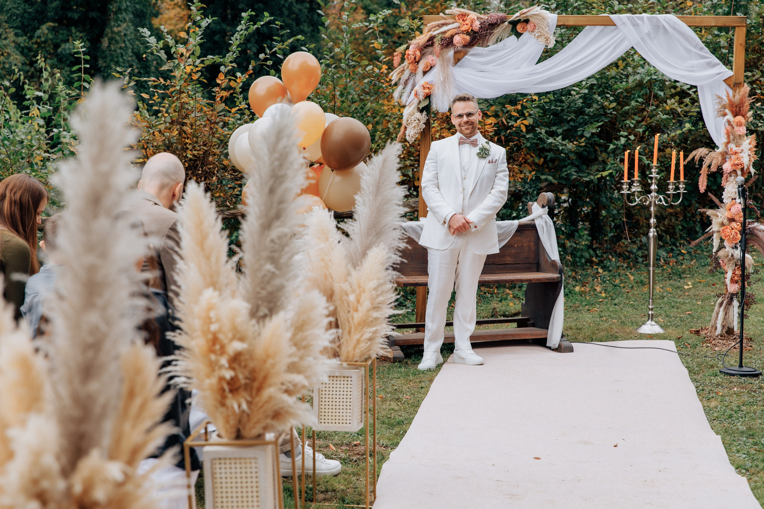 Der Bräutigam steht in einem weißen Anzug und schaut, wie seine Braut erscheint, vor dem Hintergrund einer schönen Dekoration in braun-orangenen und beigen Tönen.