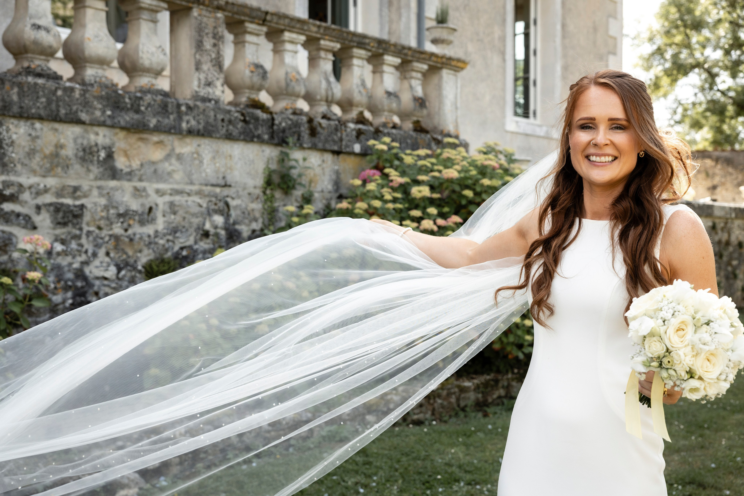 Bride outdoors with a cathedral veil trailing behind her and a white bouquet