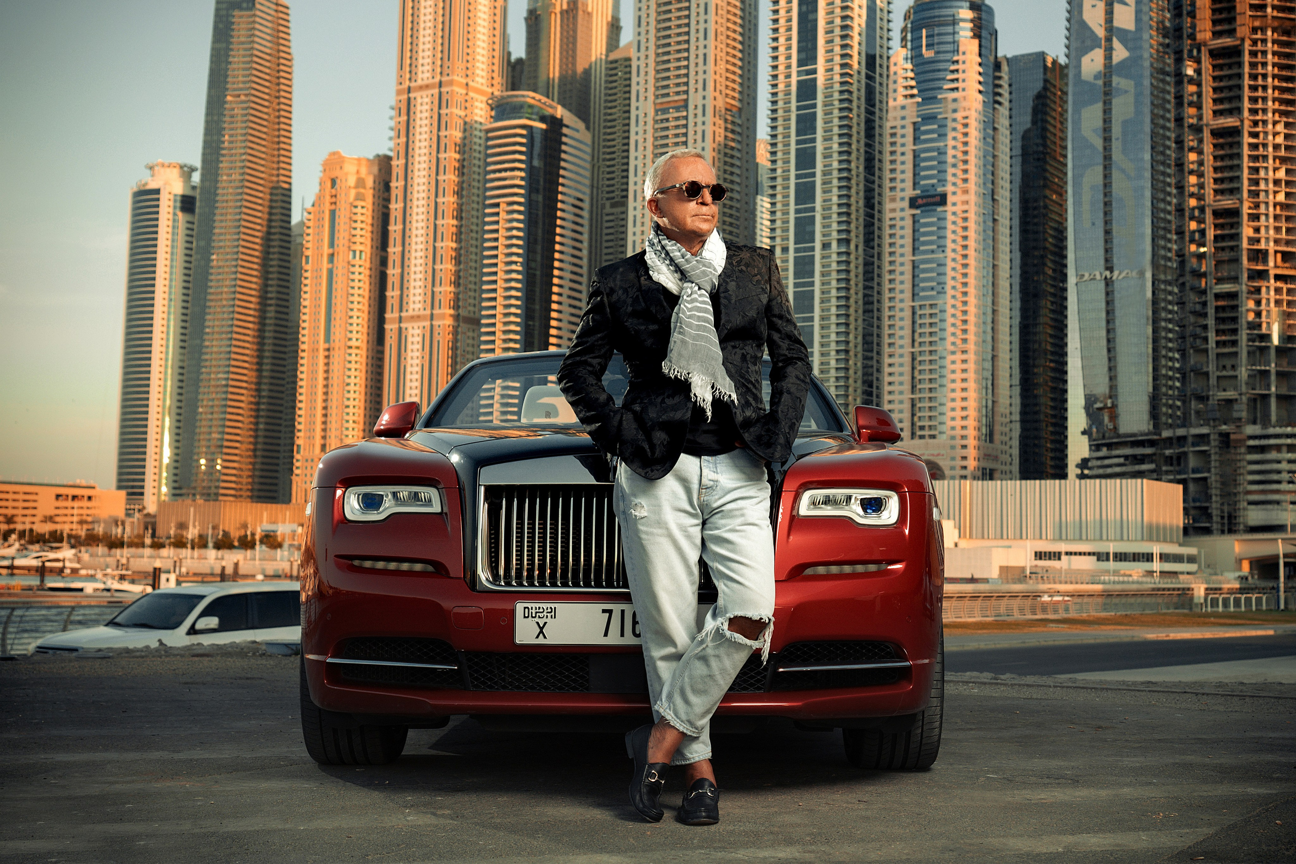 A guy stands next to a red Rolls-Royce with Dubai’s skyscrapers in the background during a beautiful sunset