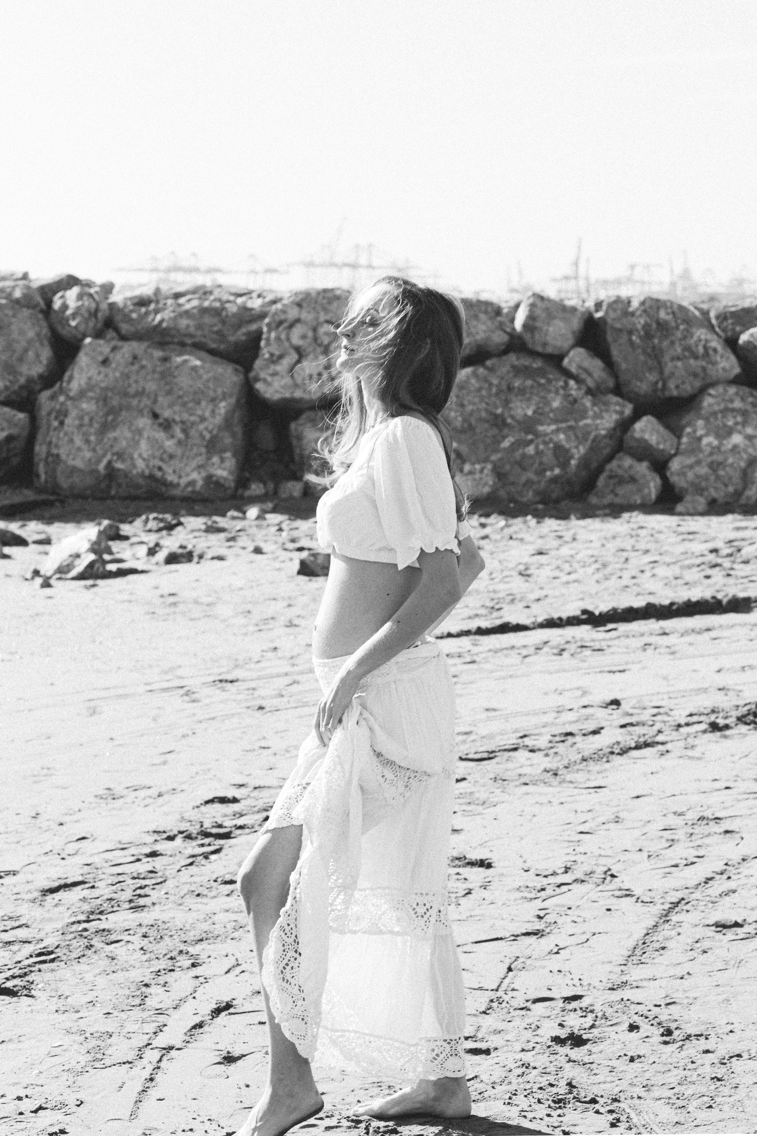 Sesión de fotos de embarazo en València, España, con una mujer embarazada caminando por la playa con un elegante vestido fluido, capturada en un estilo atemporal en blanco y negro con luz natural suave, brisa marina y una atmósfera mediterránea tranquila, resaltando la maternidad, la feminidad y la conexión con la naturaleza — ideal para fotografía de embarazo, sesiones de maternity, retratos artísticos de embarazo y sesiones de embarazo en la playa en València y en toda España.
