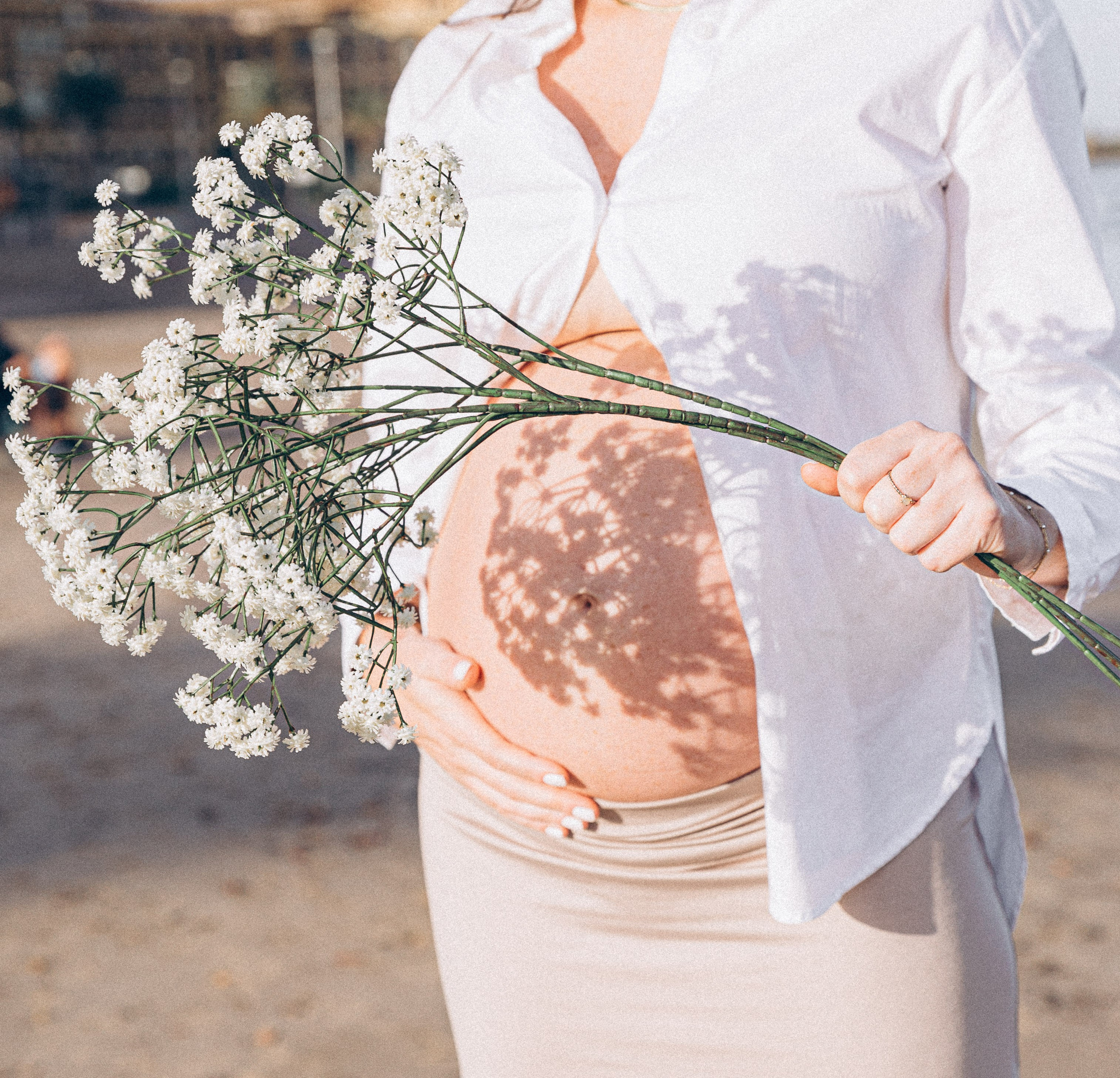 Primer plano artístico de una mujer embarazada sosteniendo flores silvestres blancas sobre su vientre, bañada por la luz cálida del sol durante una sesión de embarazo serena en la playa de Valencia, España.