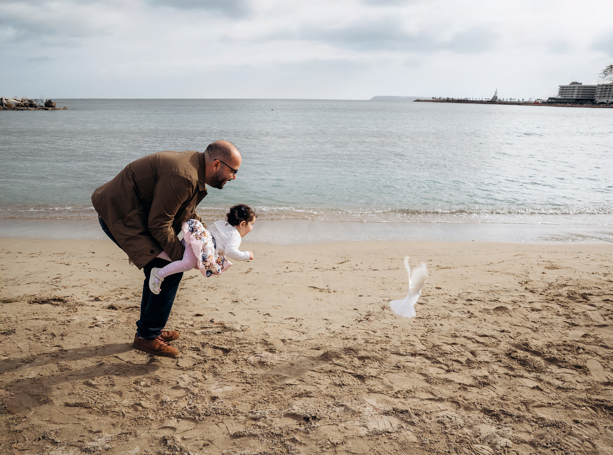 Momento divertido de un padre levantando a su hija hacia una bandada de aves en la playa de Alicante, España — una escena que capta la alegría y la curiosidad junto al mar.