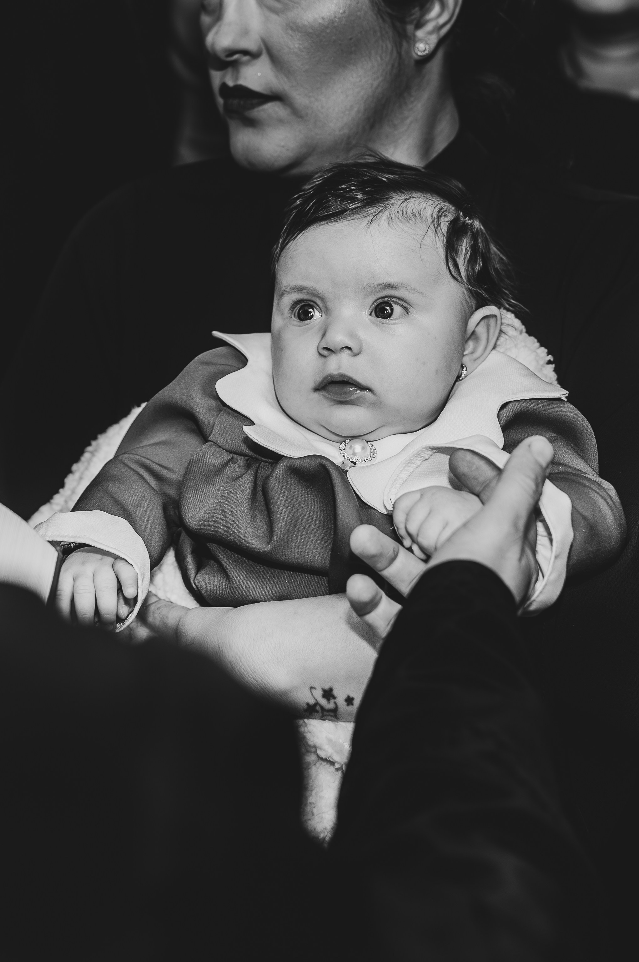 Fotografie alb-negru de impact de la botezul Evelinei Maria. Nașa ține cu grijă și tandrețe pruncul înfășat în brațe, într-un moment intim de dinaintea binecuvântării. Imagine emoționantă și atemporală.