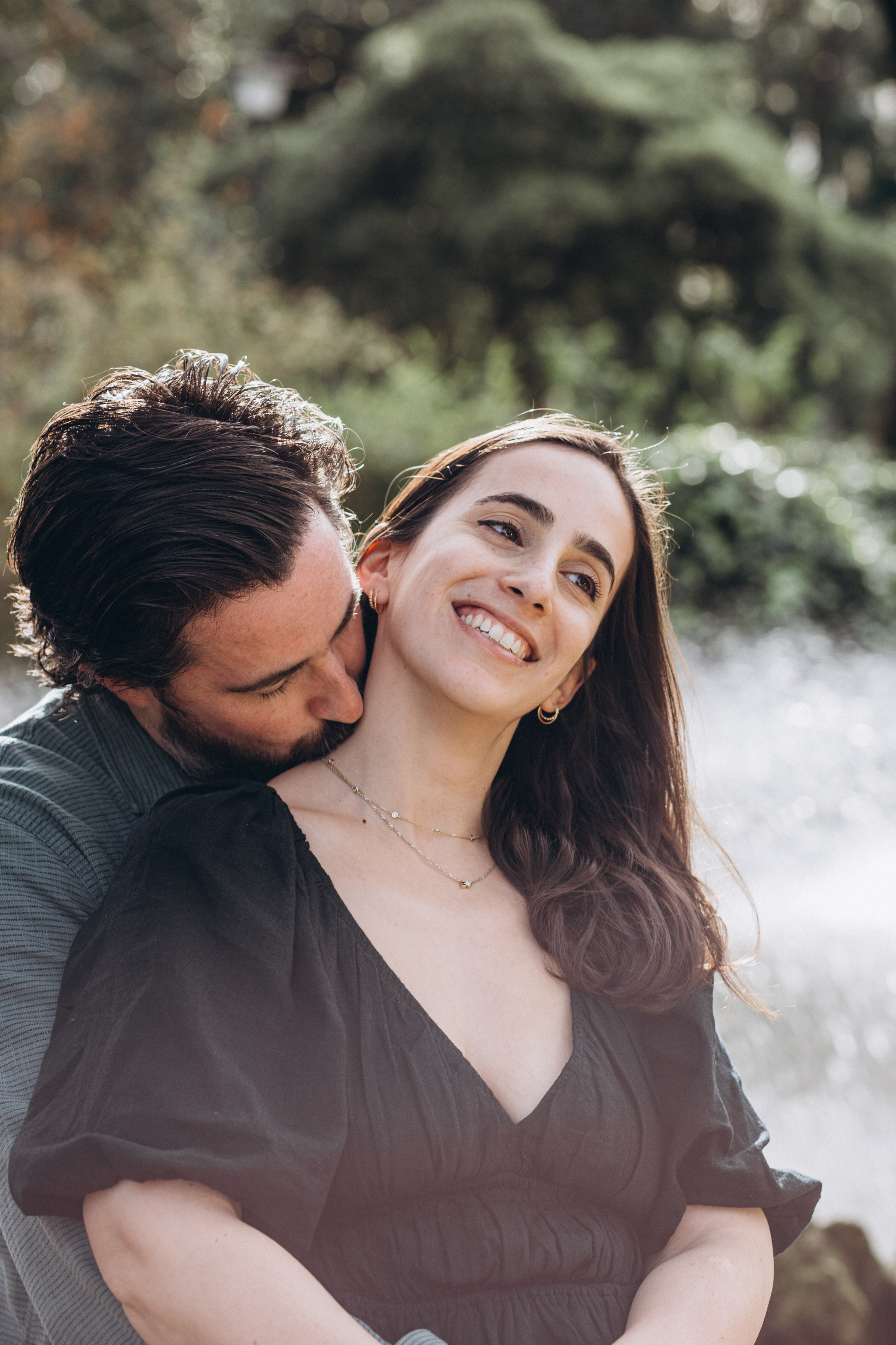 Joyful couple’s portrait of a man gently kissing his smiling partner’s neck from behind, surrounded by lush greenery and soft natural light, capturing a tender and affectionate moment Valencia, Spain.