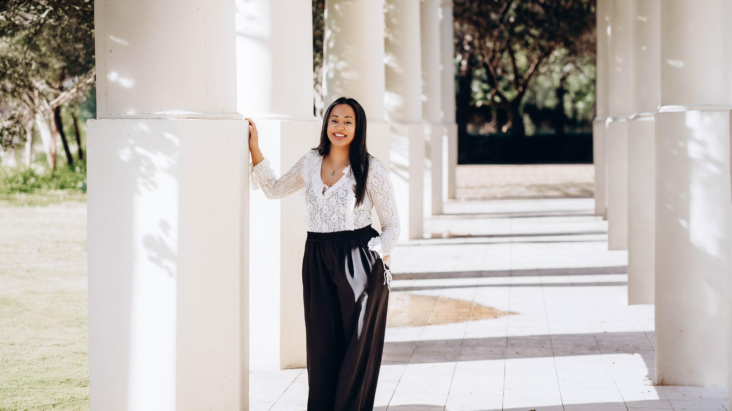 Женская портретная фотосессия в Валенсии, Испания: улыбающаяся женщина стоит у белых архитектурных колонн в мягком естественном свете, в стильном образе, сочетающем casual и chic — идеально для лайфстайл и уличной портретной съемки.