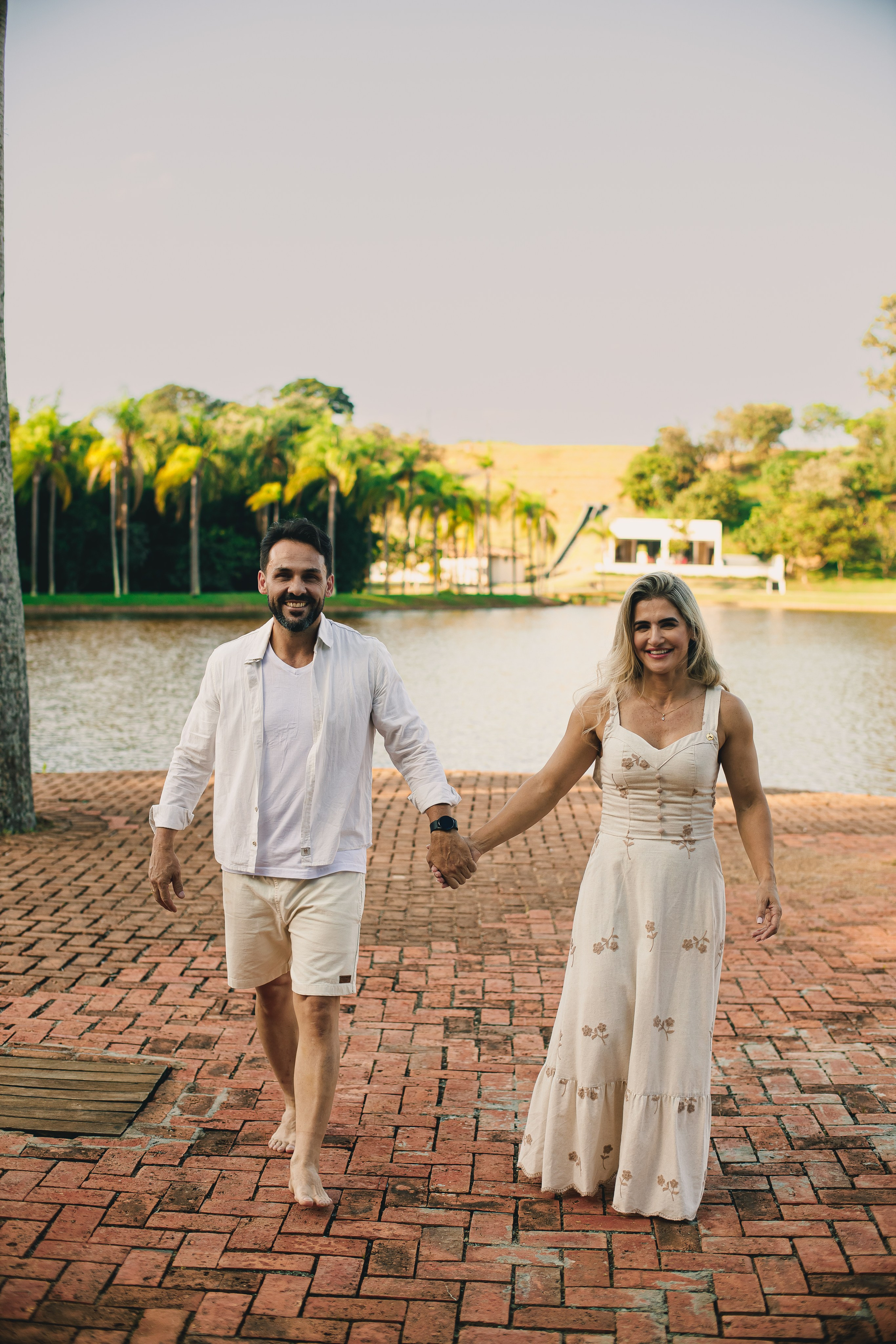 Casal sorrindo e caminhando de mãos dadas no calçadão de pedra do Resort das Oliveiras, com lago e palmeiras ao fundo