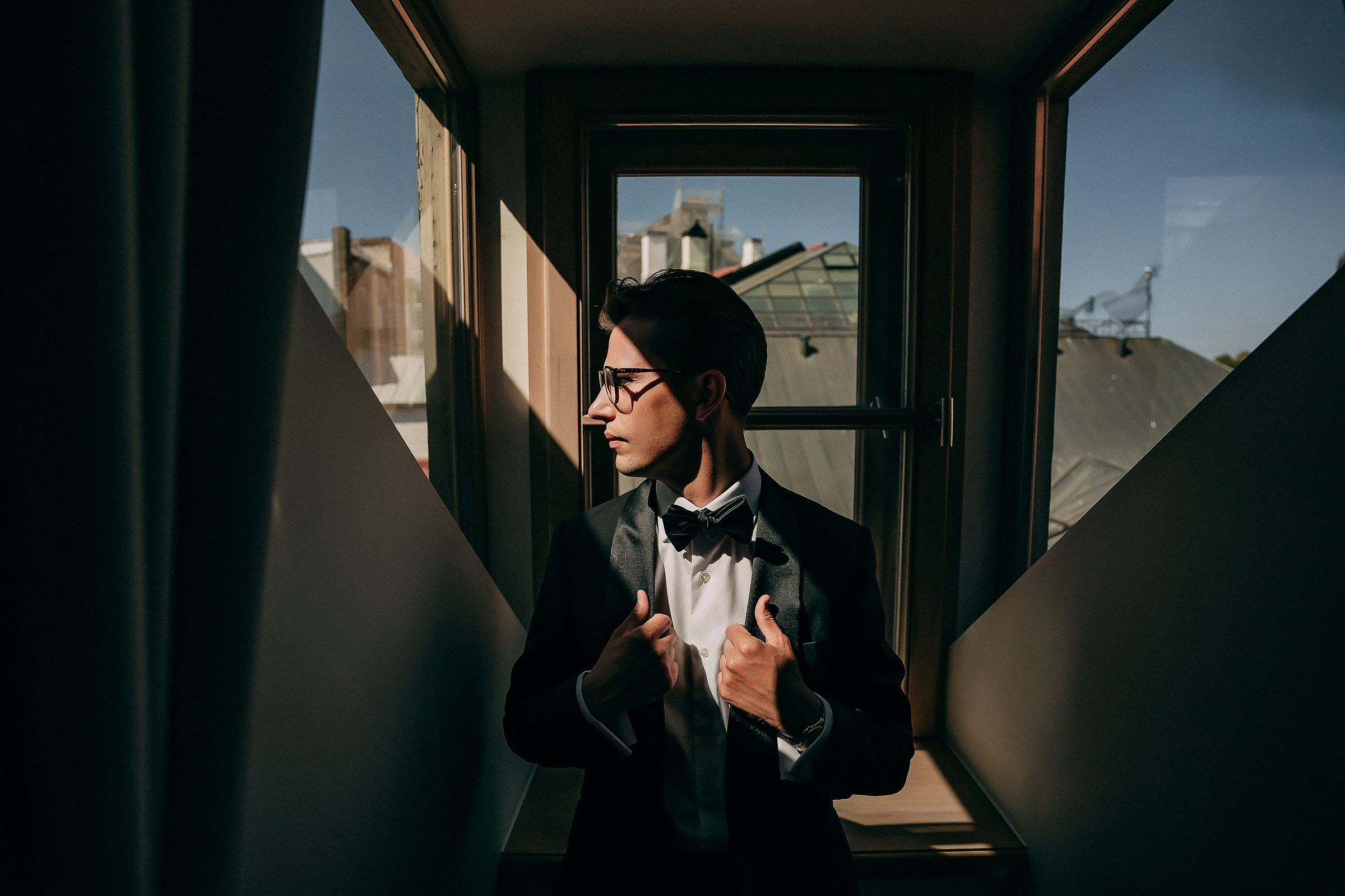 The groom in a tuxedo with a bow tie stands in front of a window with a view of rooftops and a clear sky.The lighting creates dramatic shadows and highlights on the suit and surrounding walls.