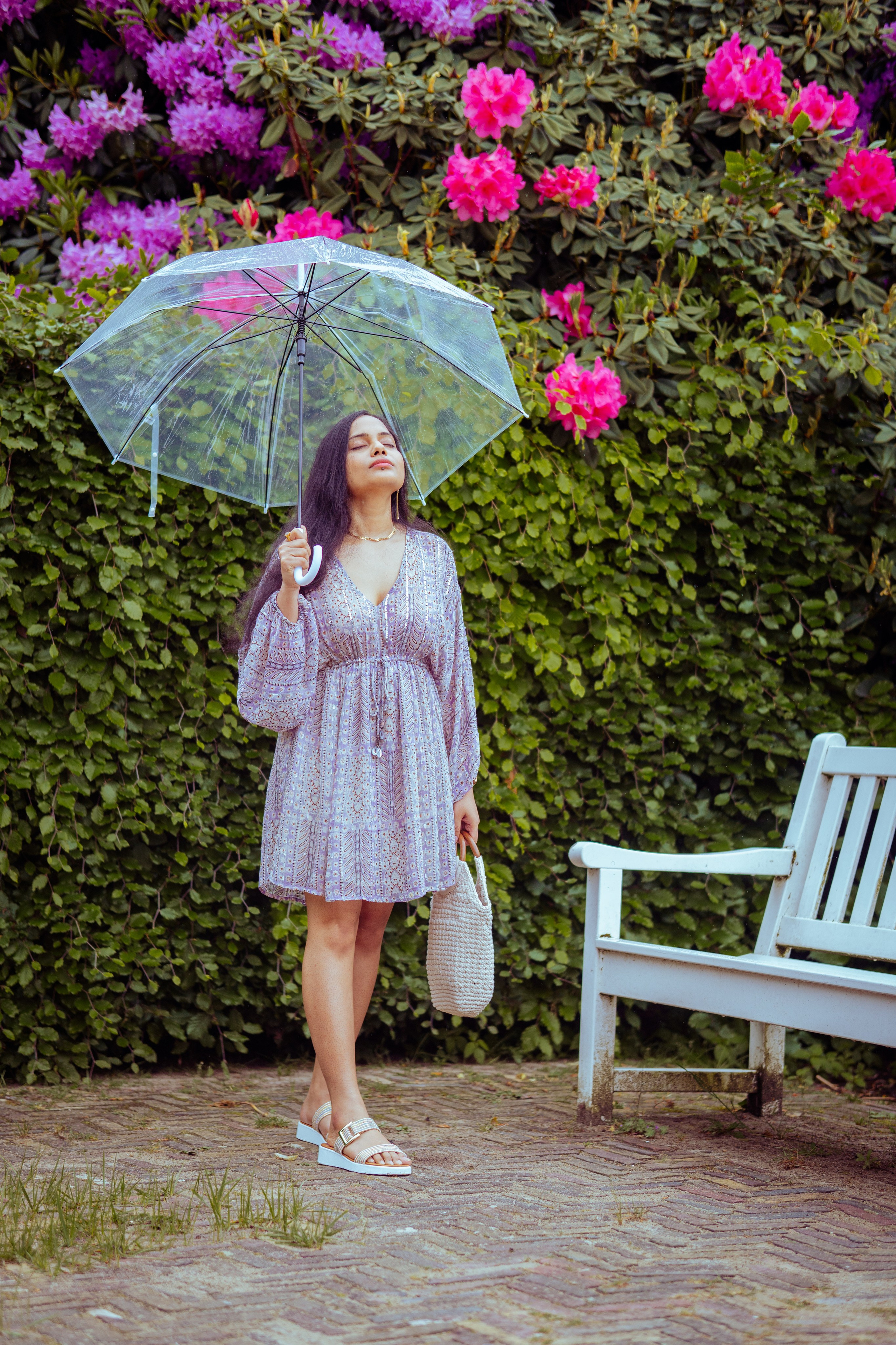 woman standing near a white bench with flowers as background and holding a transparent umbrella