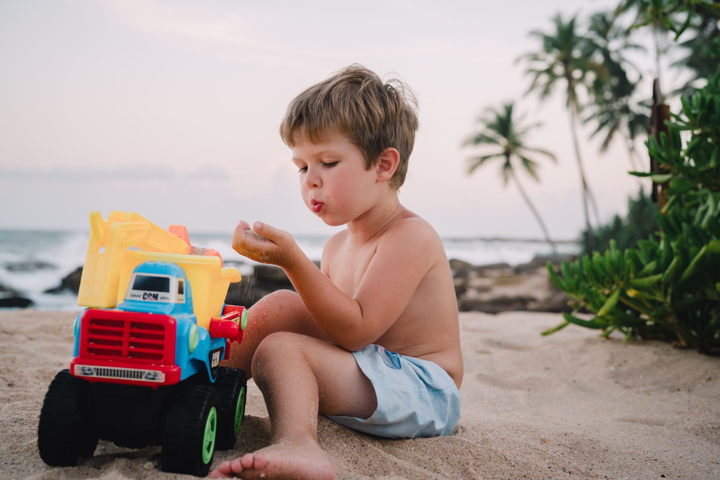 The boy playing with a car on a beach with palm trees on a background