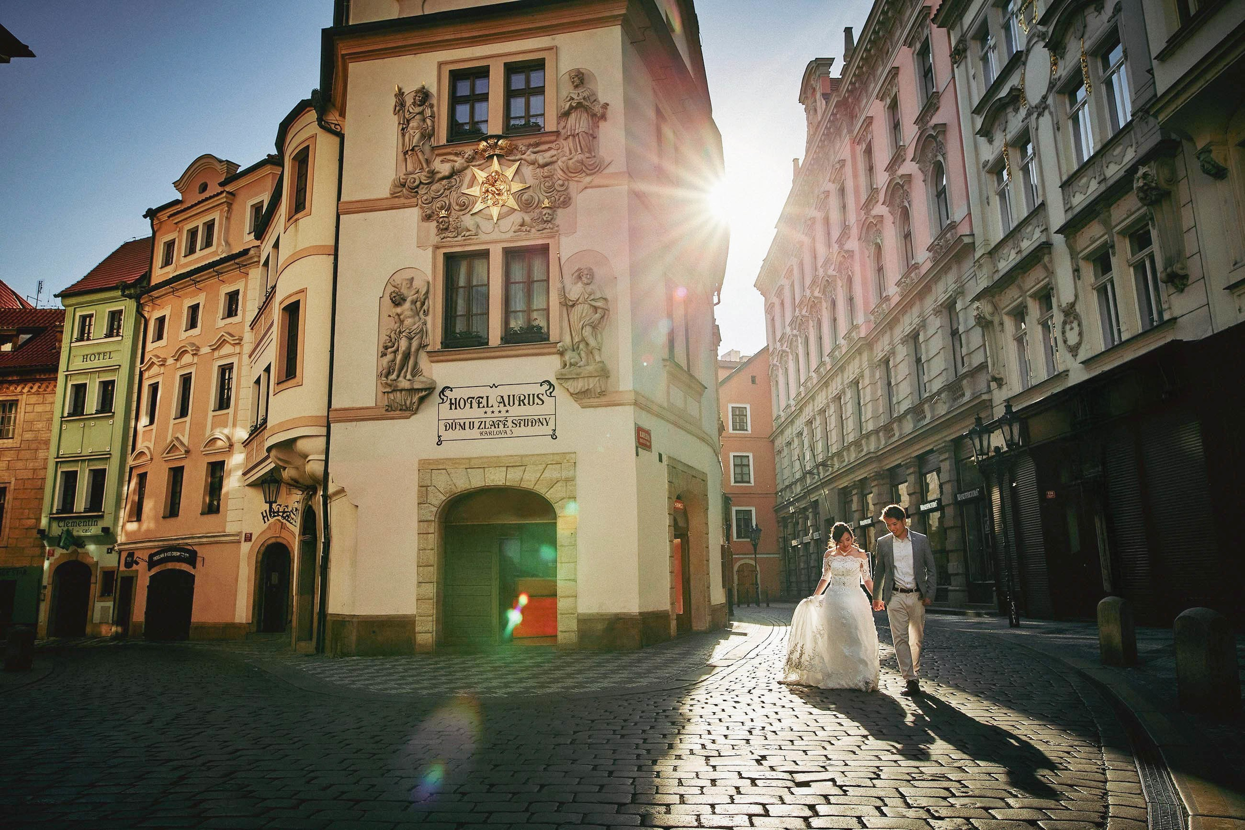 A burst of sun flare between historical buidings in Prague illuminates the way as a young Asian bride and groom walk along the cobblestone 