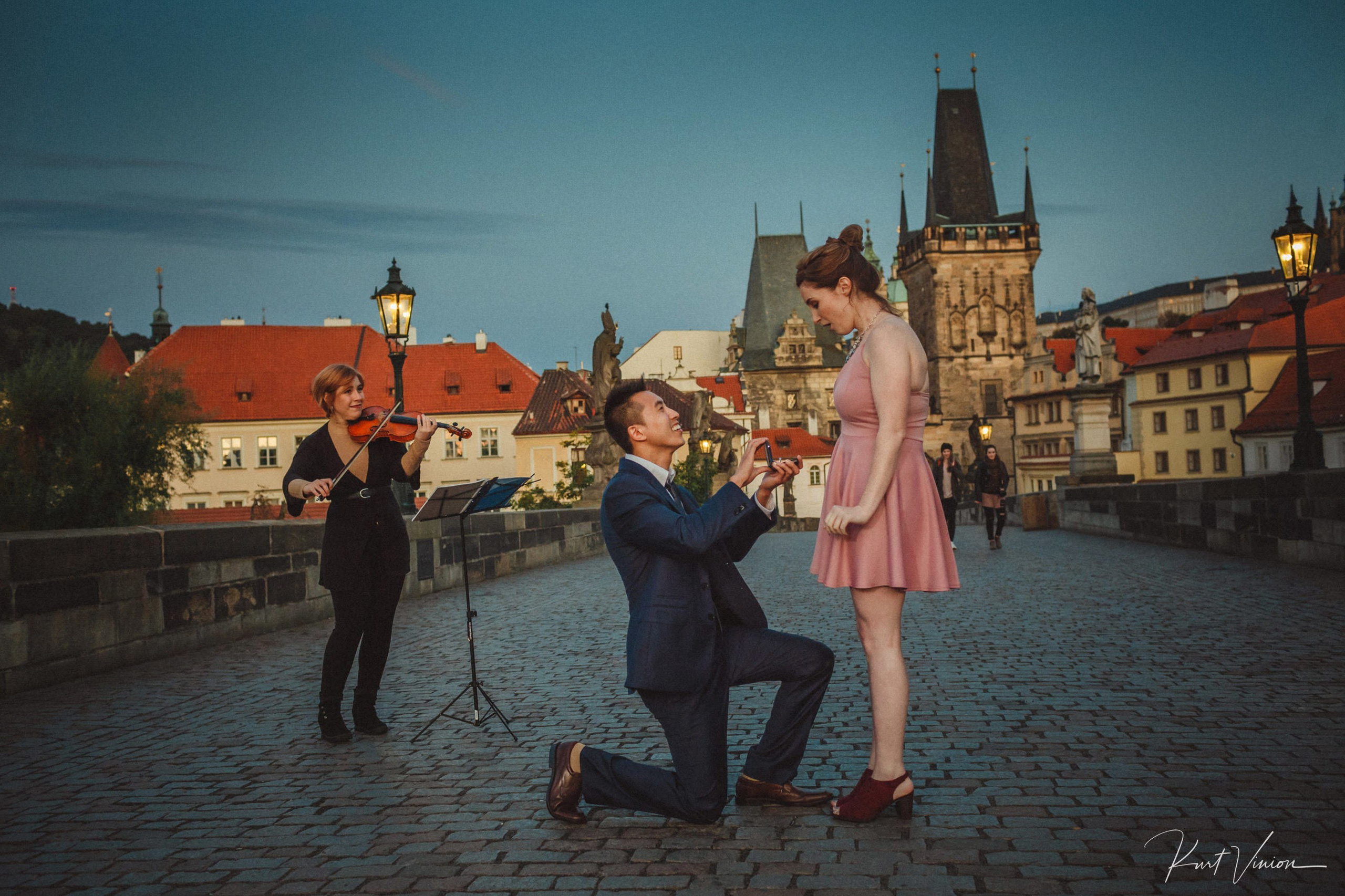 Woman in pink dress shocked as man proposes on knee with violinist performing on Charles Bridge at dawn.