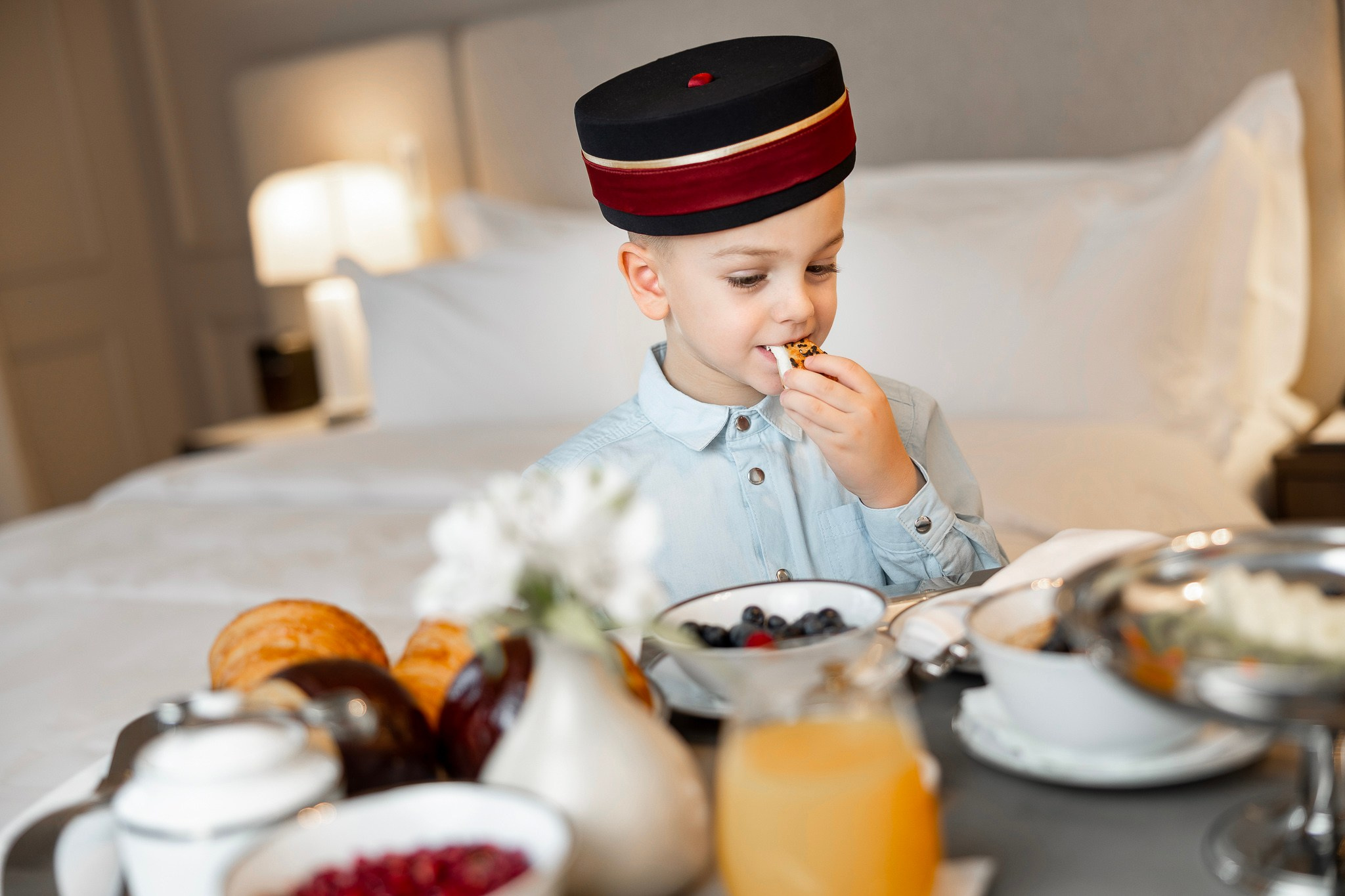 decak sa kapom od vratara hotela sedi na krevetu hotelske sobe i jede raznovrsni dorucak
