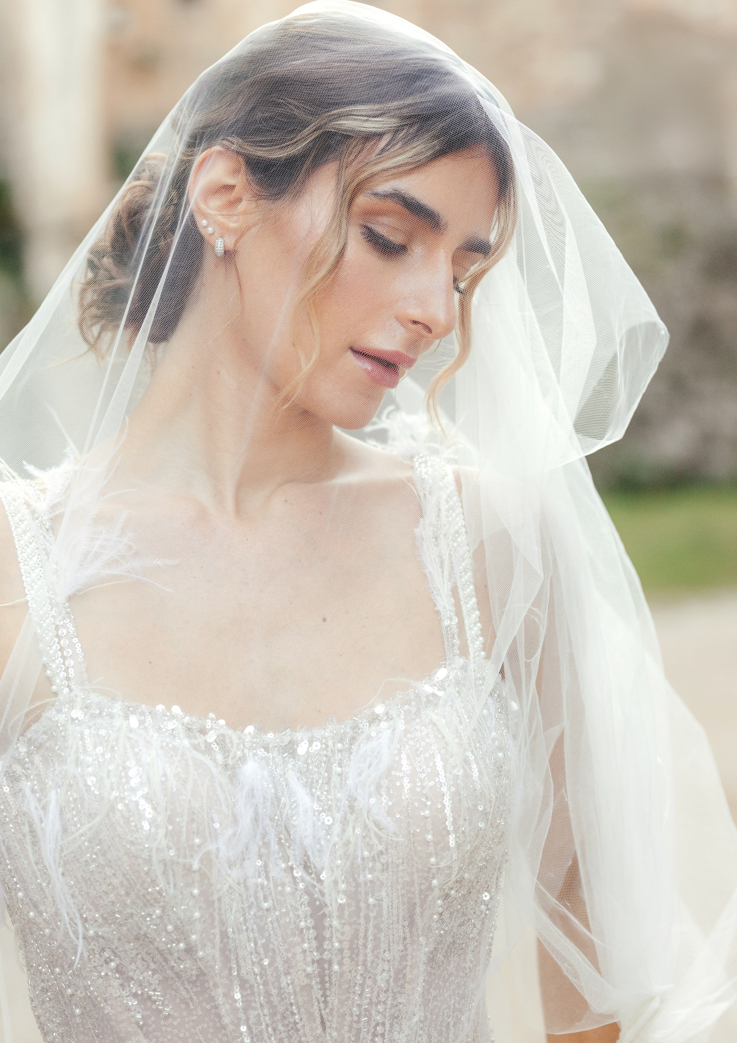 A radiant bride stands in the narrow, sunlit streets of Rhodes' Old Town, her intricate lace wedding dress contrasting beautifully with the weathered stone walls behind her. The editorial-style portrait highlights her serene expression and the timeless elegance of the historic setting, with soft natural light accentuating her features.