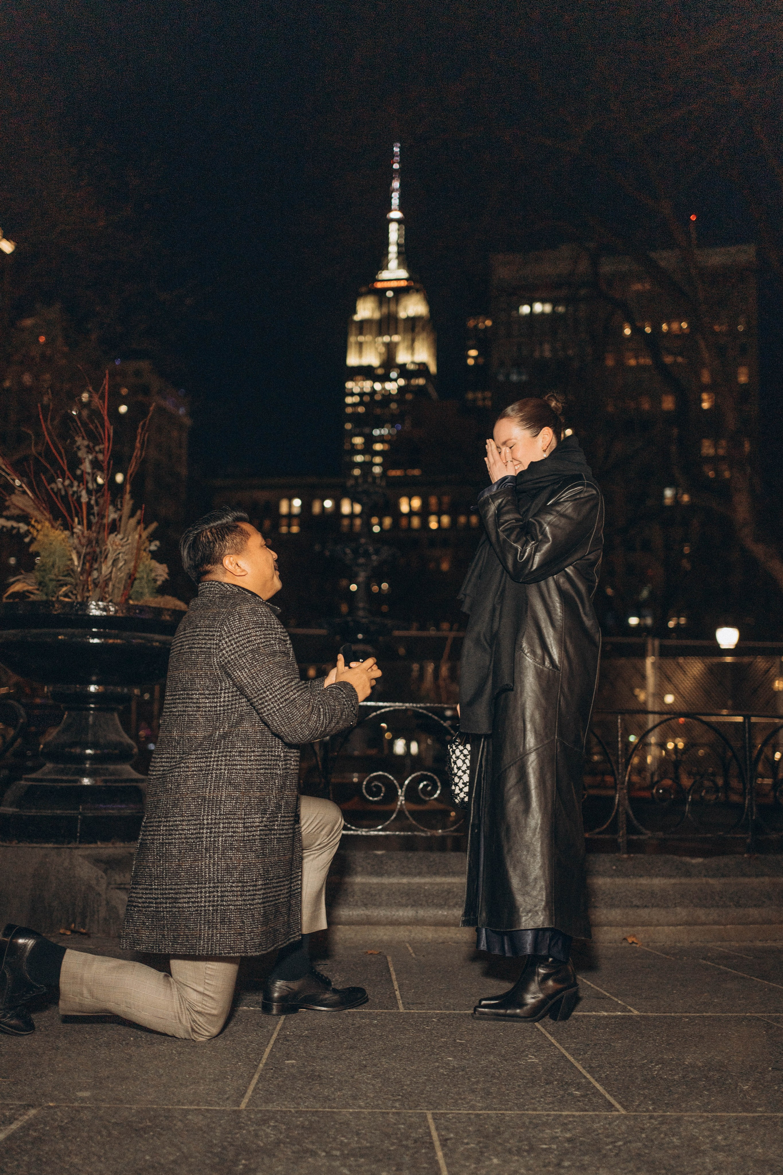 Surprise proposal by Bow Bridge in Central Park.Man kneeling with skyline view in Brooklyn.