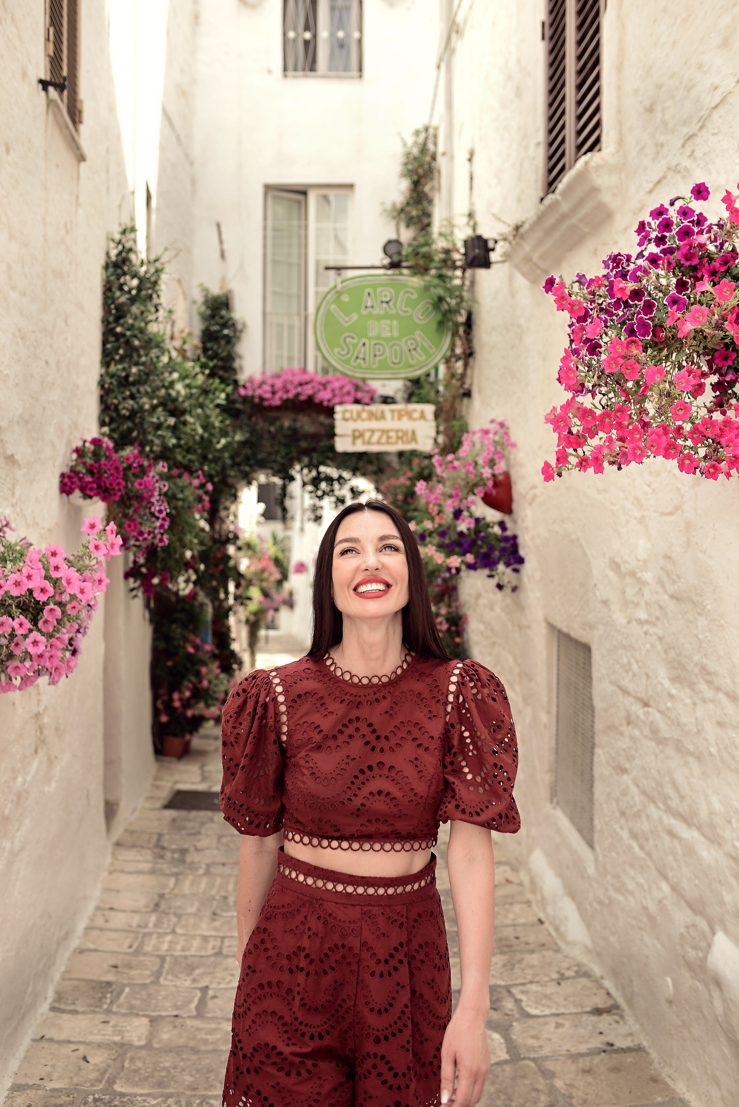 Smiling woman in a burgundy two-piece outfit walks down a charming alley of Ostuni filled with pink and violet flowers in bloom, under a restaurant sign.