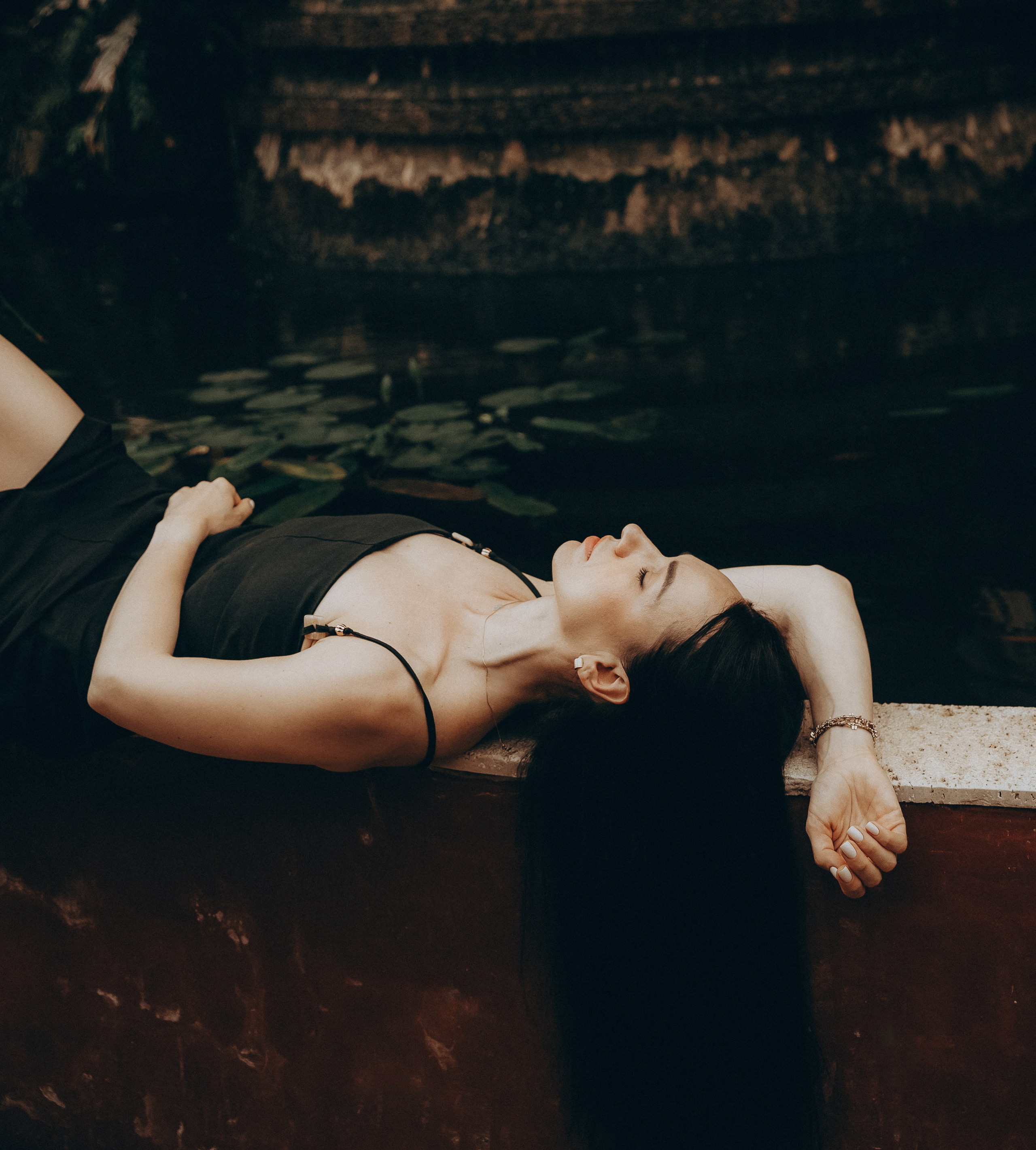 Женщина в черном платье спокойно лежит у пруда с лилиями, отражающего мягкий золотой свет, снято в Малаге, Испания — художественная и умиротворяющая портретная фотография, подчеркивающая природную красоту и моменты покоя, идеально подходит для портретных фотосессий в Малаге и Испании.