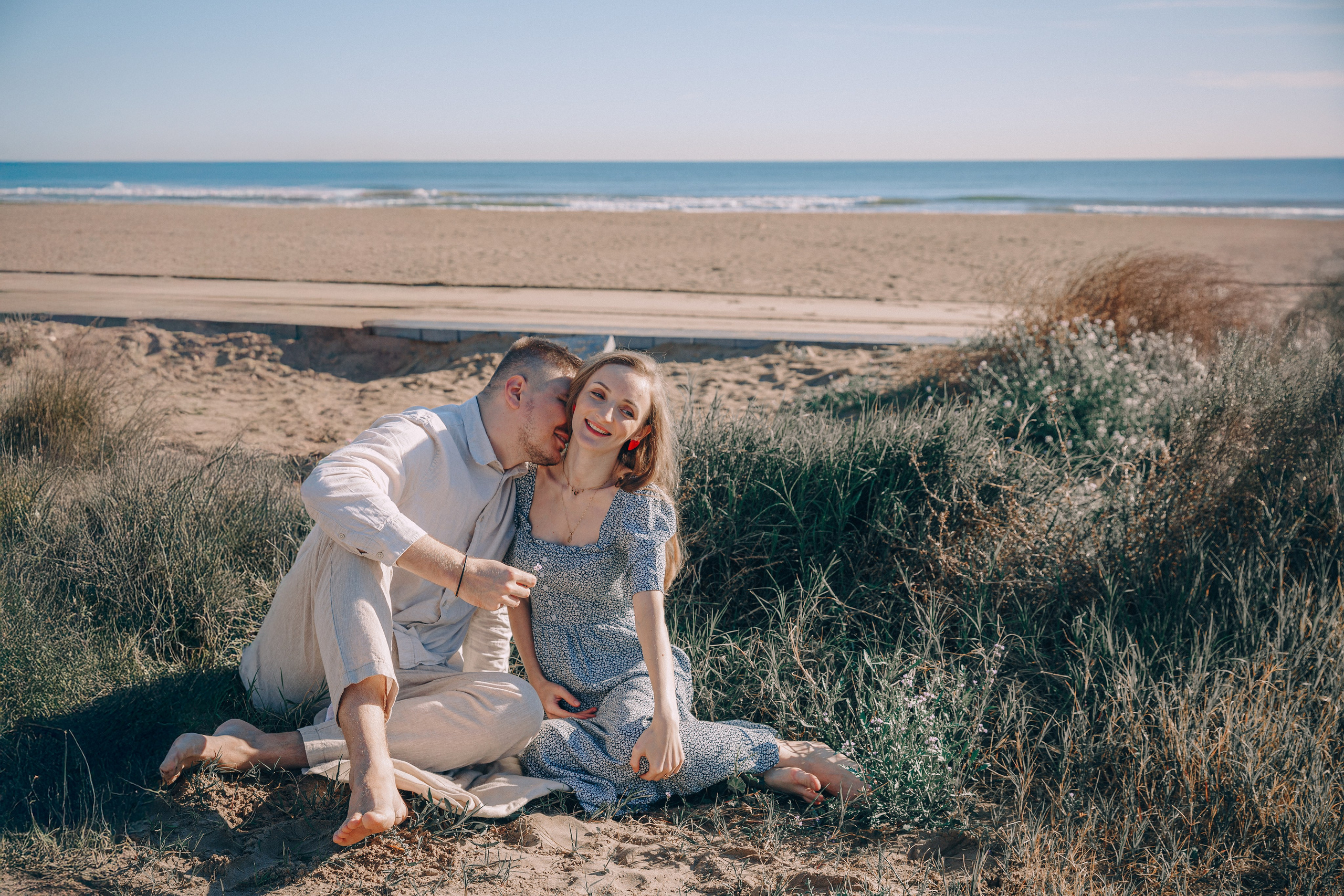 Sesión de fotos love story romántica de pareja en València, España, con una pareja sentada sobre dunas de arena junto a la playa mediterránea, compartiendo un momento íntimo y delicado con el mar de fondo — fotografía natural de pareja, sesión love story en la playa, retratos estilo compromiso y sesiones emocionales de pareja en València y en toda España.