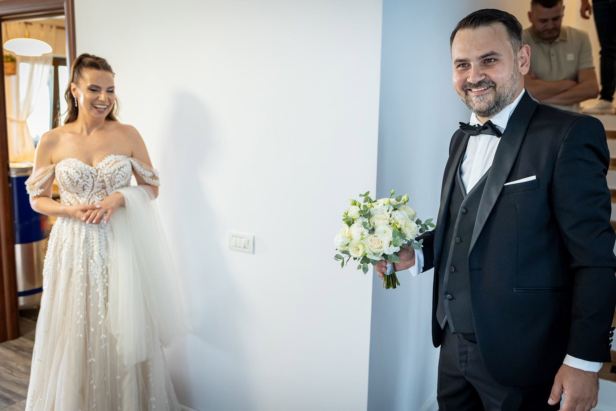 Moment first look la nuntă, mirele cu buchetul de trandafiri albi zâmbind către mireasă în rochie brodată