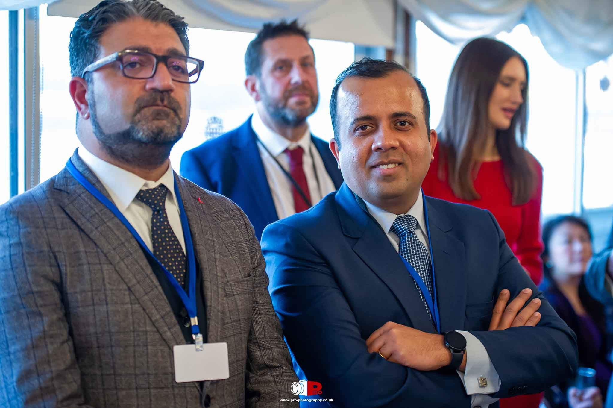 Two professional men in business suits smiling at a corporate networking event with a group of attendees in the background.