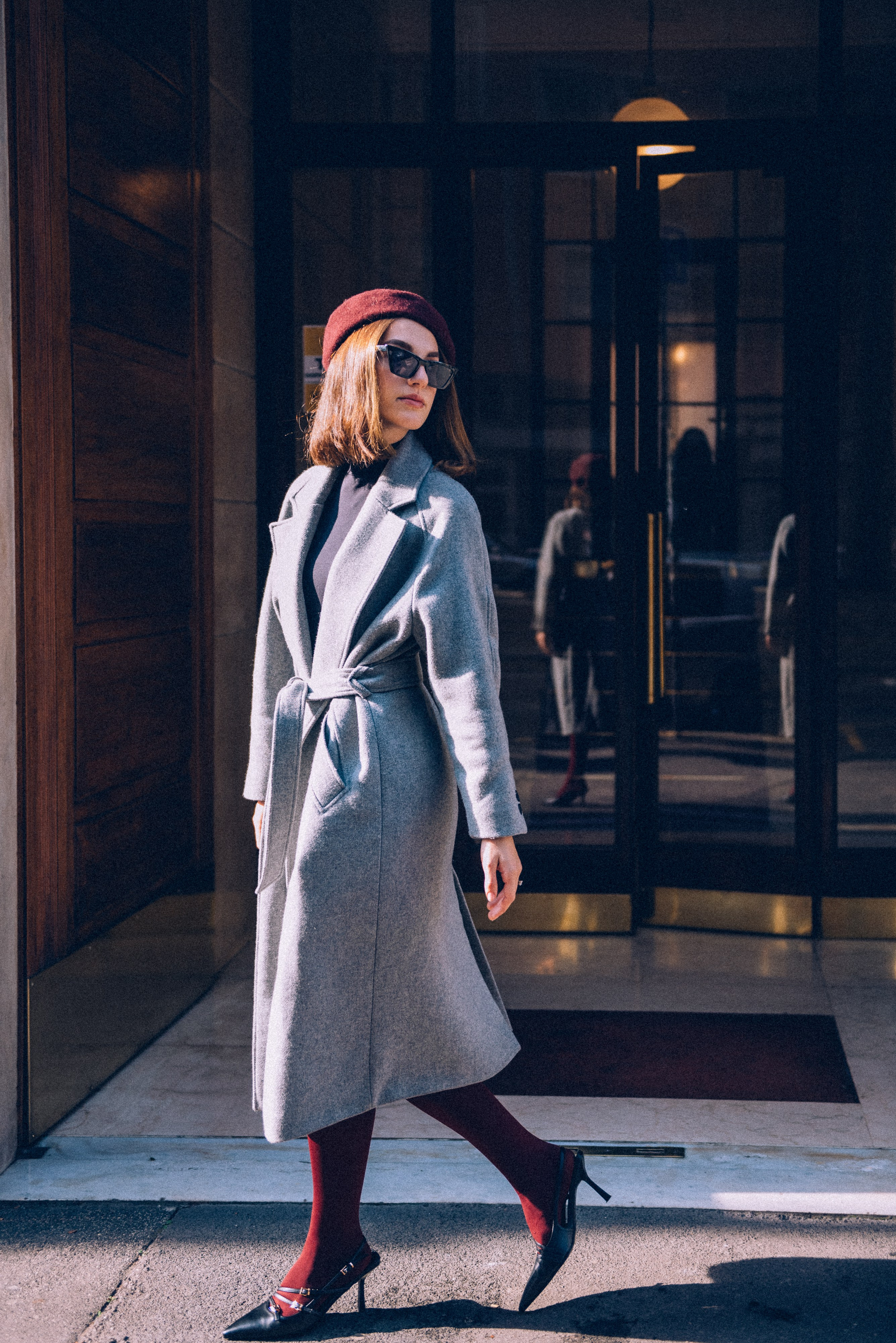 Fashionable woman in a gray coat and burgundy tights stepping confidently into the sunlight outside a classic Milanese building, showcasing elegant street style. Natural light photographer Milan.