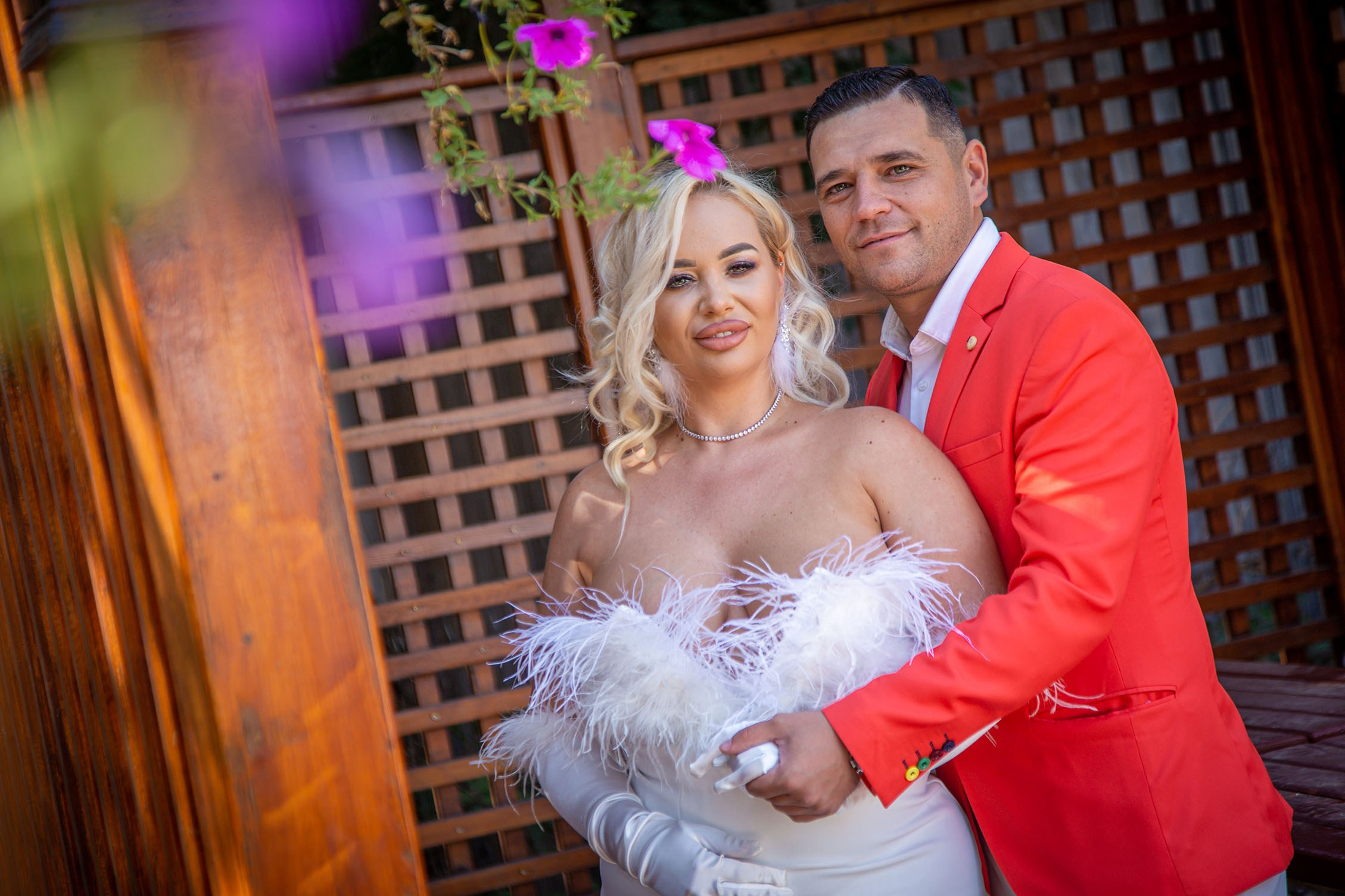 Mireasă blondă în rochie albă cu boa din pene, alături de mire în sacou roșu, ședință foto de nuntă romantică în aer liber realizată de EduardFoto