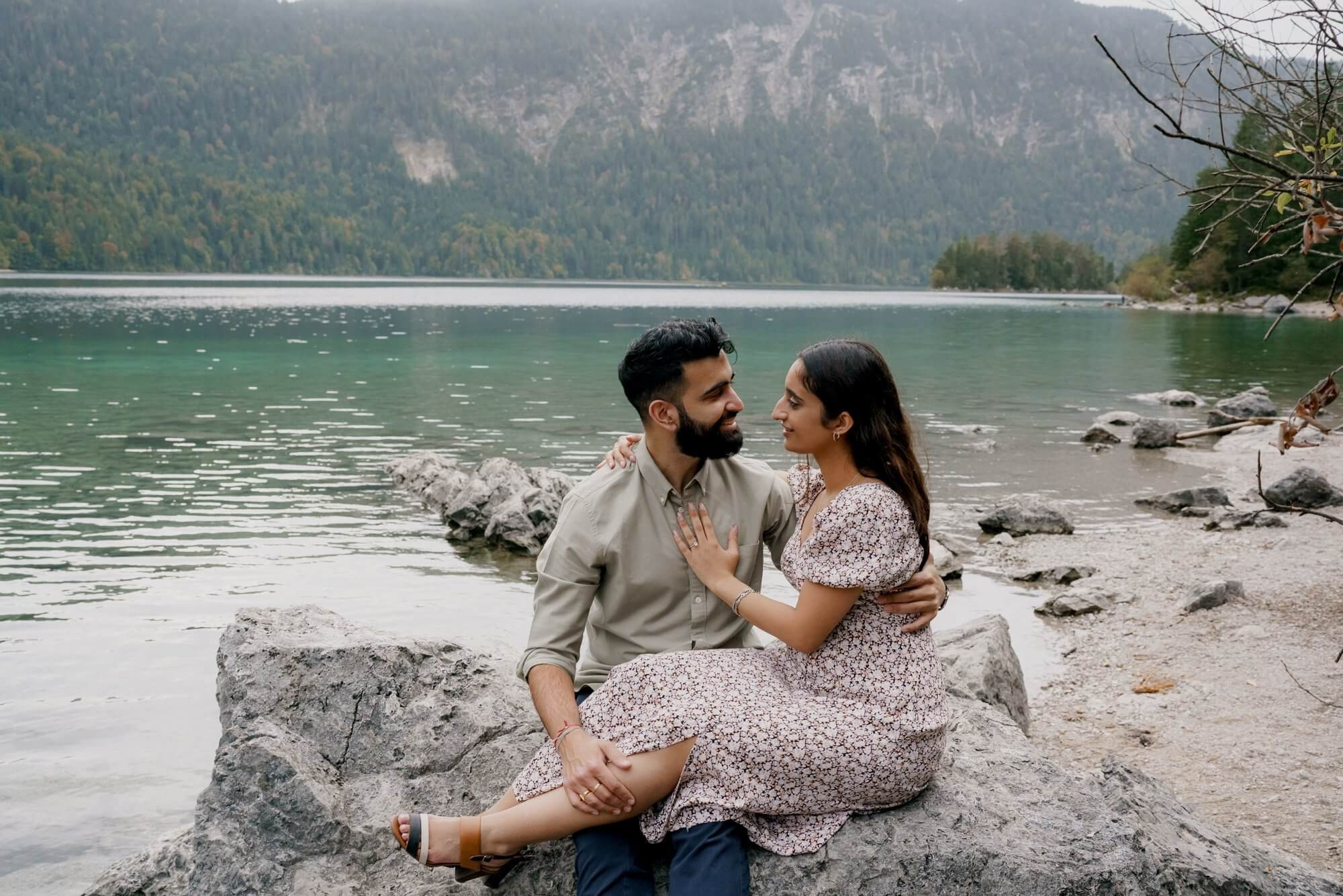 Couple sitting on driftwood at the shore of Eibsee lake with turquoise water and forest during engagement session Germany