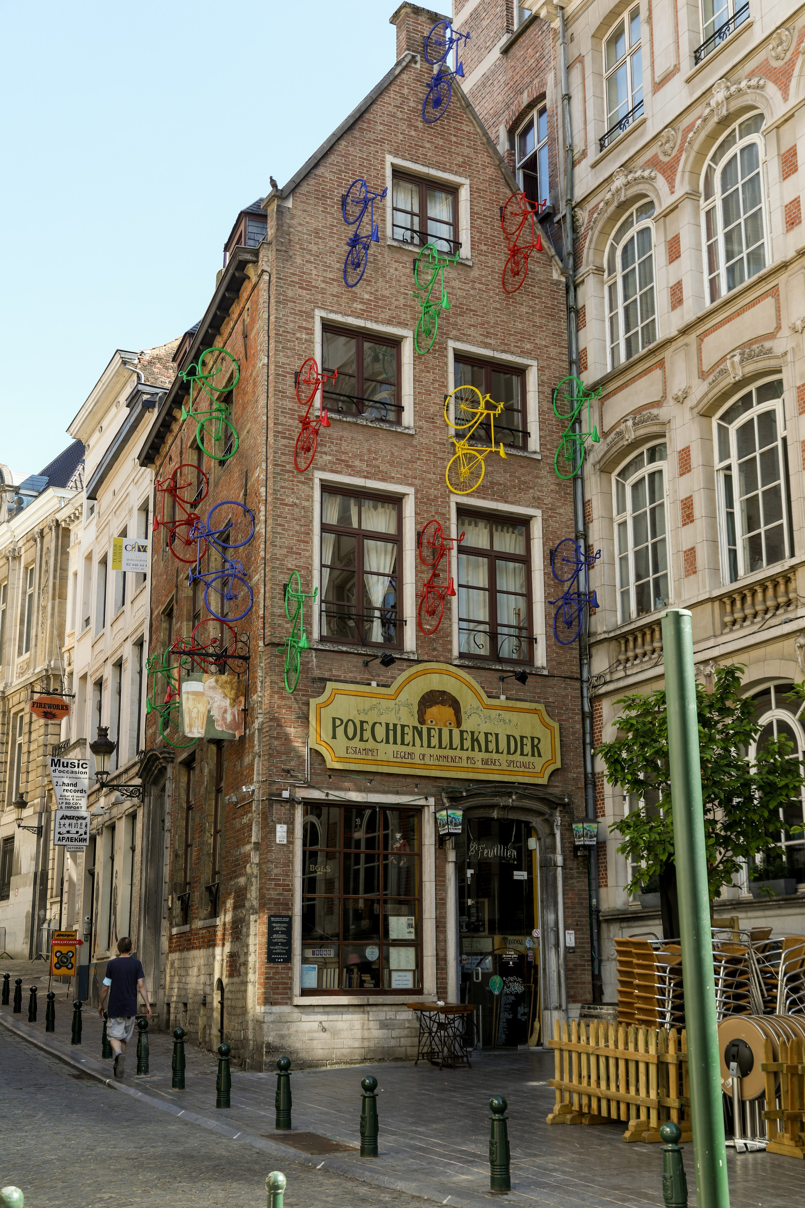 The exterior facade of the historic Poechenellekelder pub in Brussels, a brick building decorated with many colorful red, green, and yellow bicycles. Outdoor seating with wooden tables and chairs is set up on the cobblestone street.