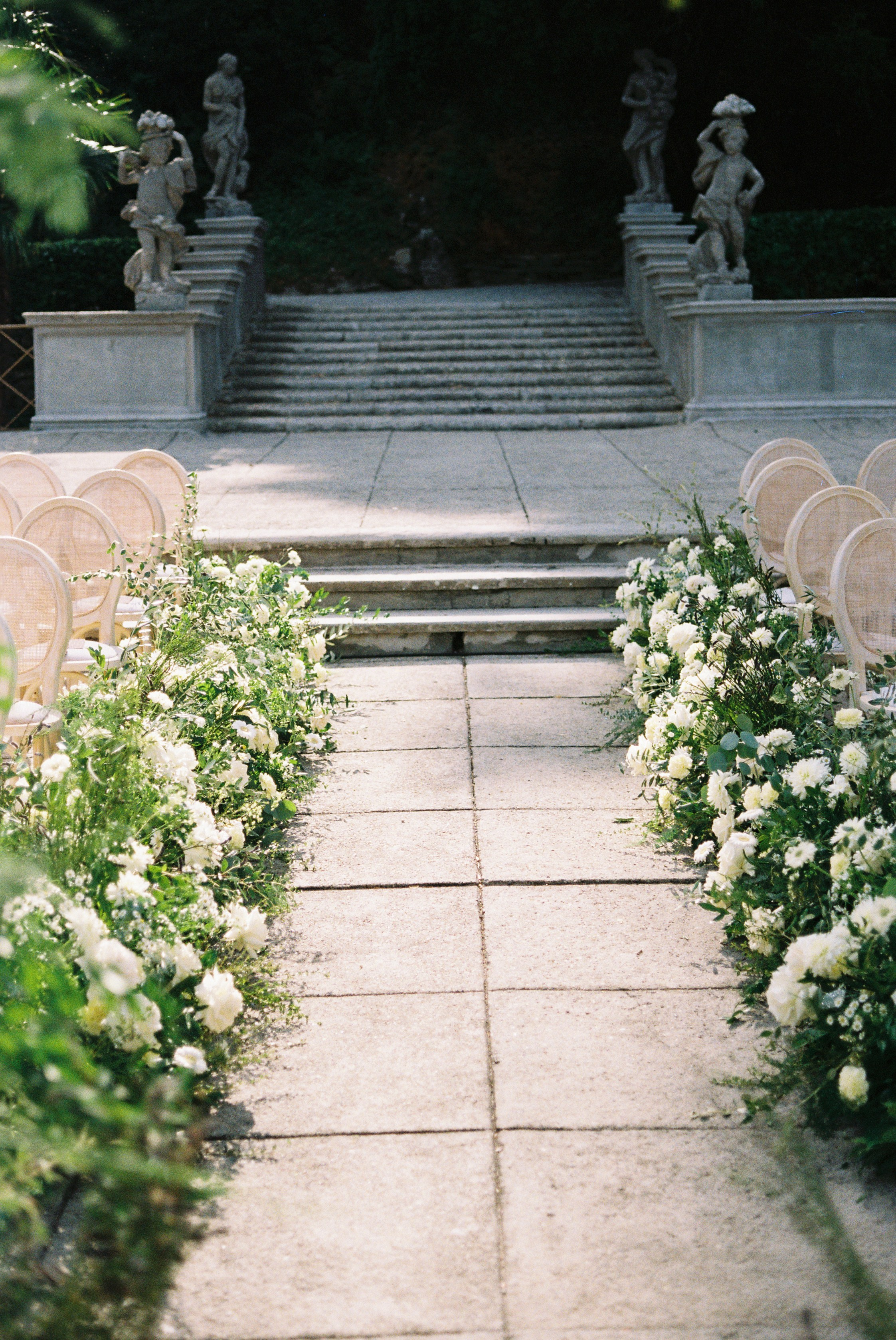 Wedding aisle lined with white flowers, leading to stone steps flanked by classical statues.