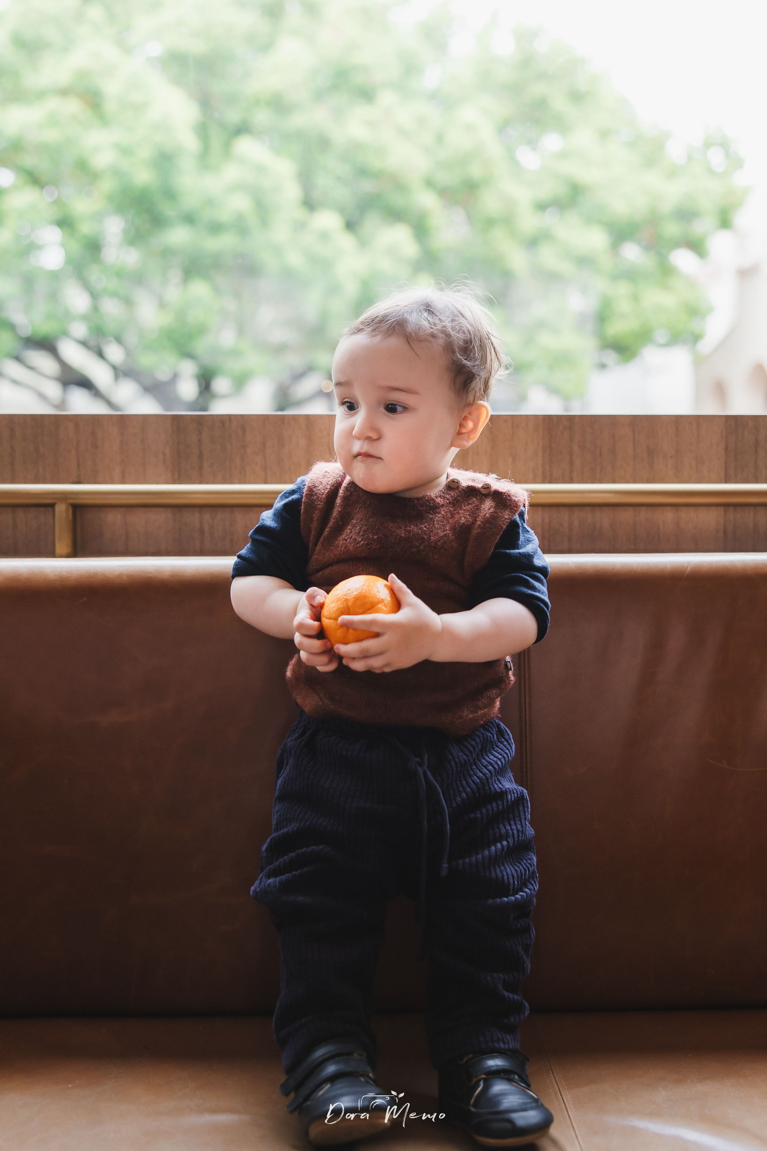 In this at-home photo, the baby is standing by the window, playing with an orange.