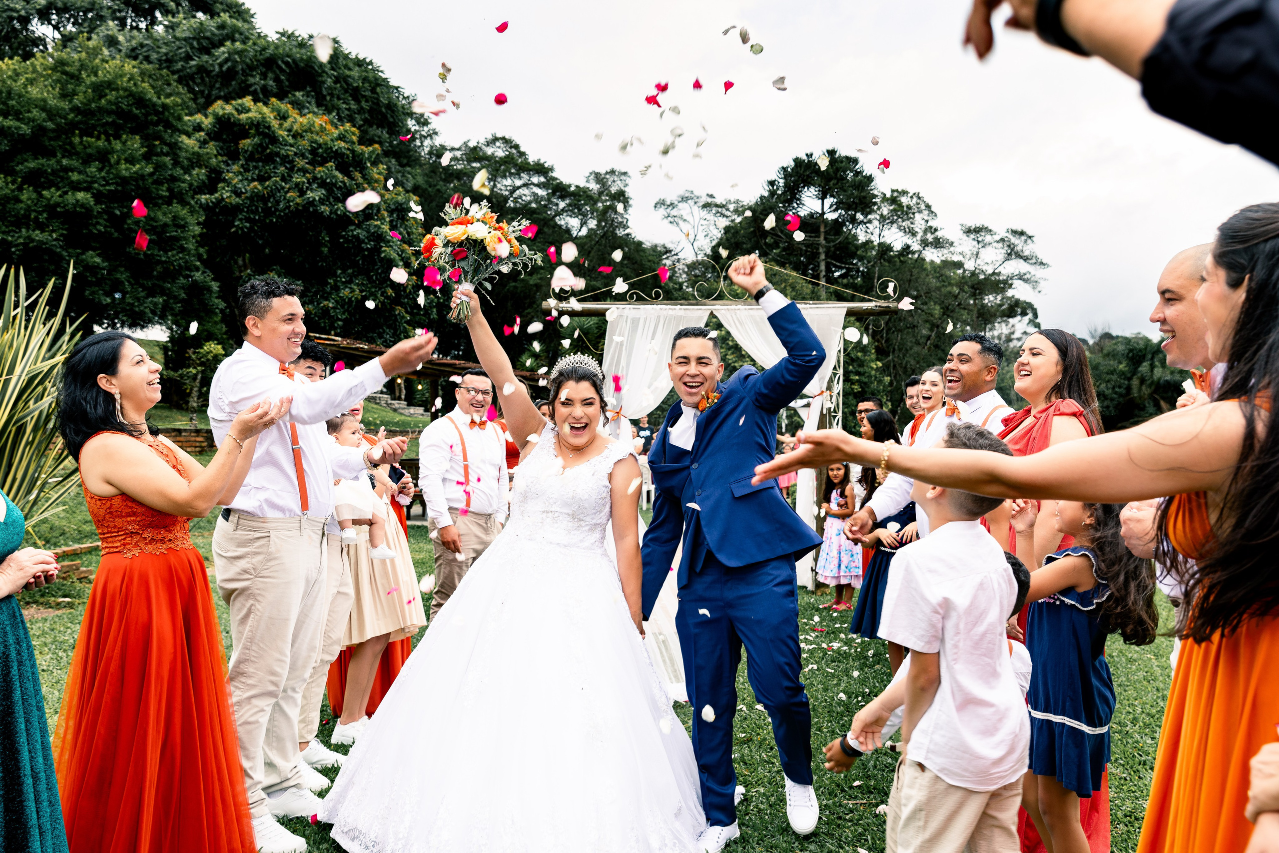 Noivos comemorando com os braços erguidos sob chuva de pétalas, cercados por amigos e familiares em festa.