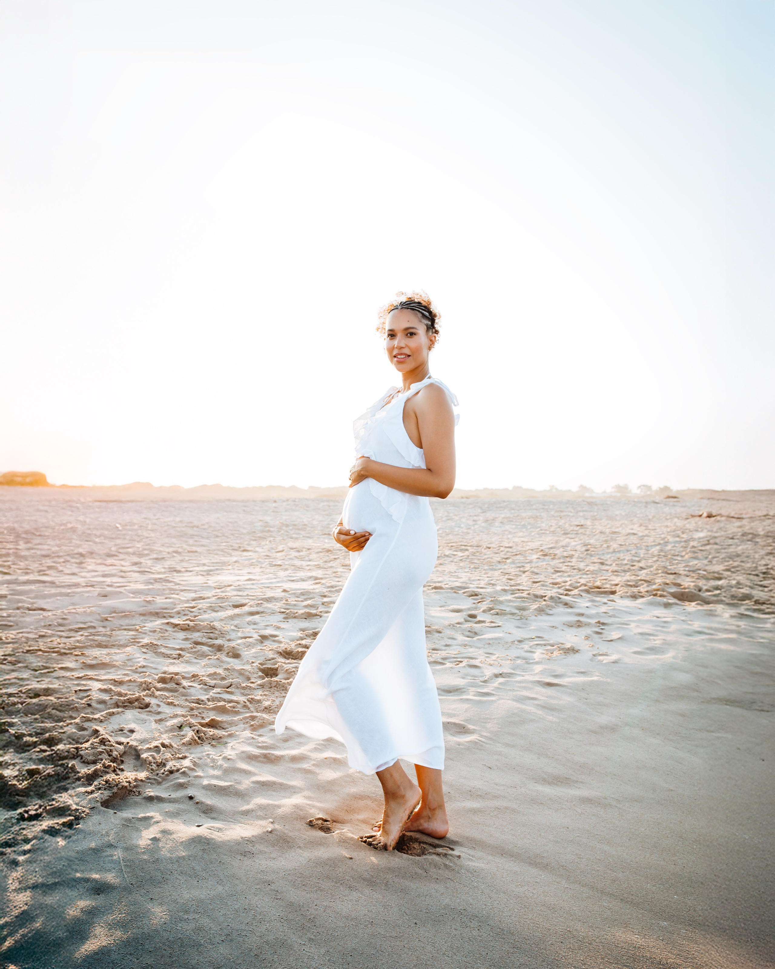 Sesión de fotos de embarazo al atardecer en la playa de Alicante, España — futura mamá radiante con vestido blanco fluido posando descalza sobre la arena. Inspiración perfecta para sesiones naturales y elegantes de embarazo en Alicante y la Costa Blanca.