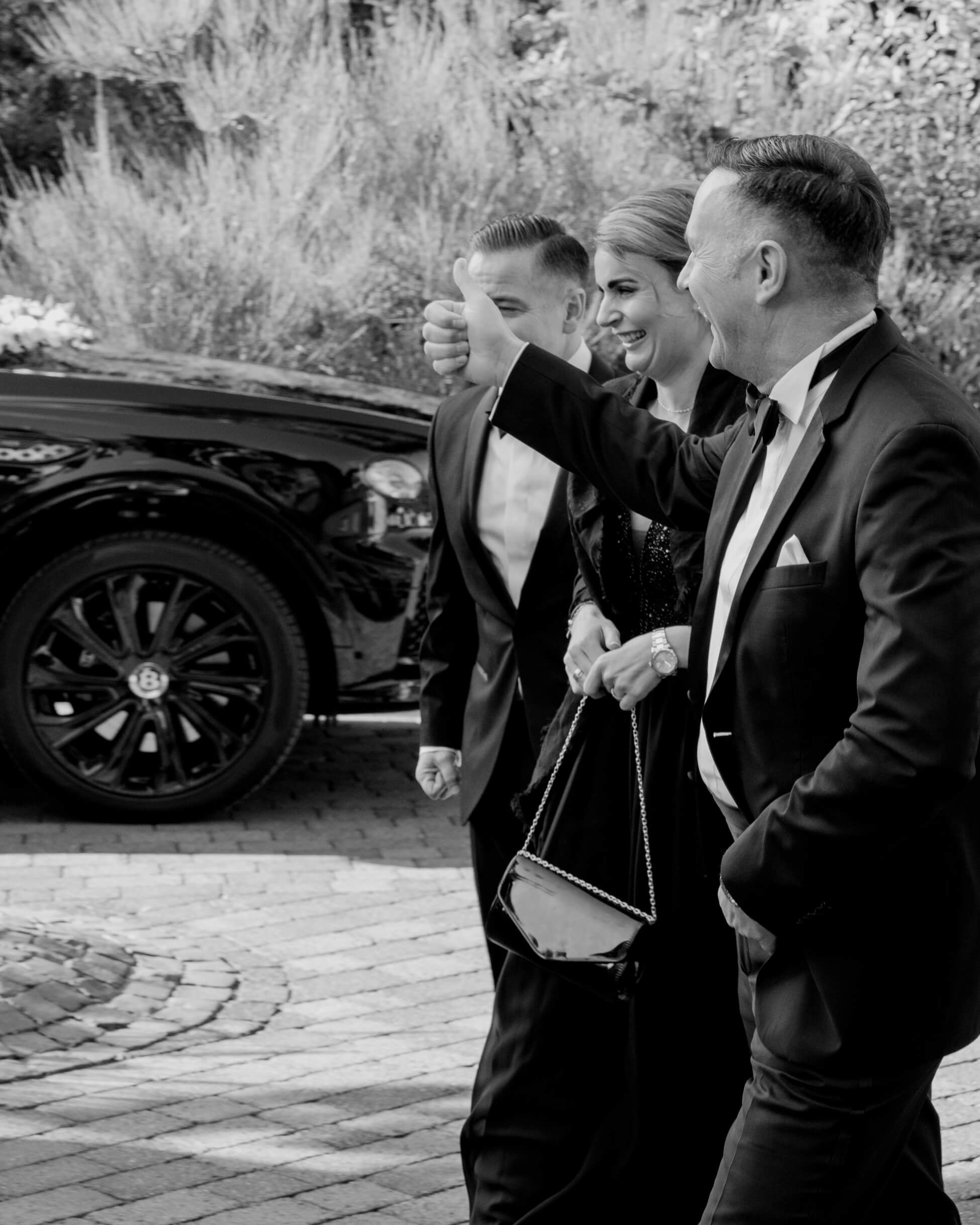 Two men in black tuxedos beside luxury Bentley, happy to see the bride coming, black tie wedding Frankfurt Villa Rothschild