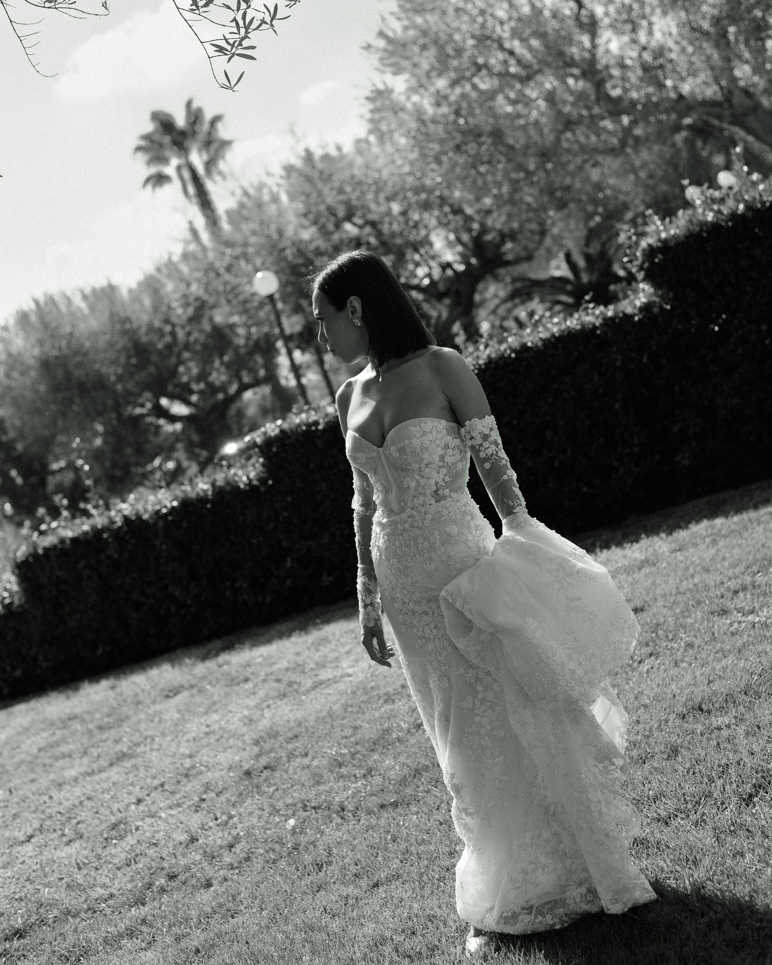 Black and white photo of a bride standing in a garden holding the train of her wedding dress