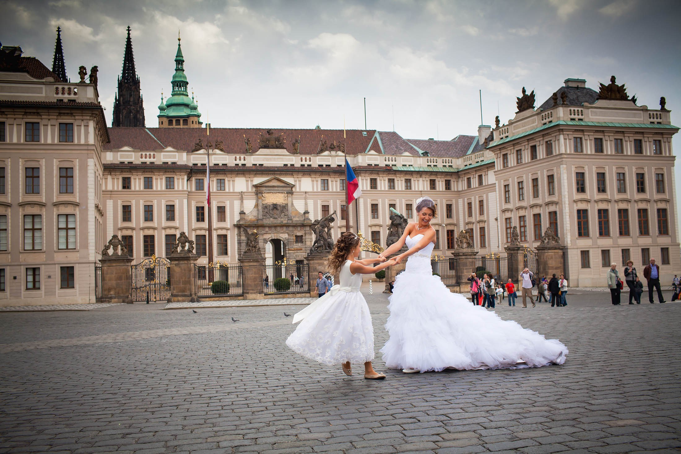 A bride in a stylish wedding dress holds her young daughter's hand as they spin in circles in front of Prague Castle.