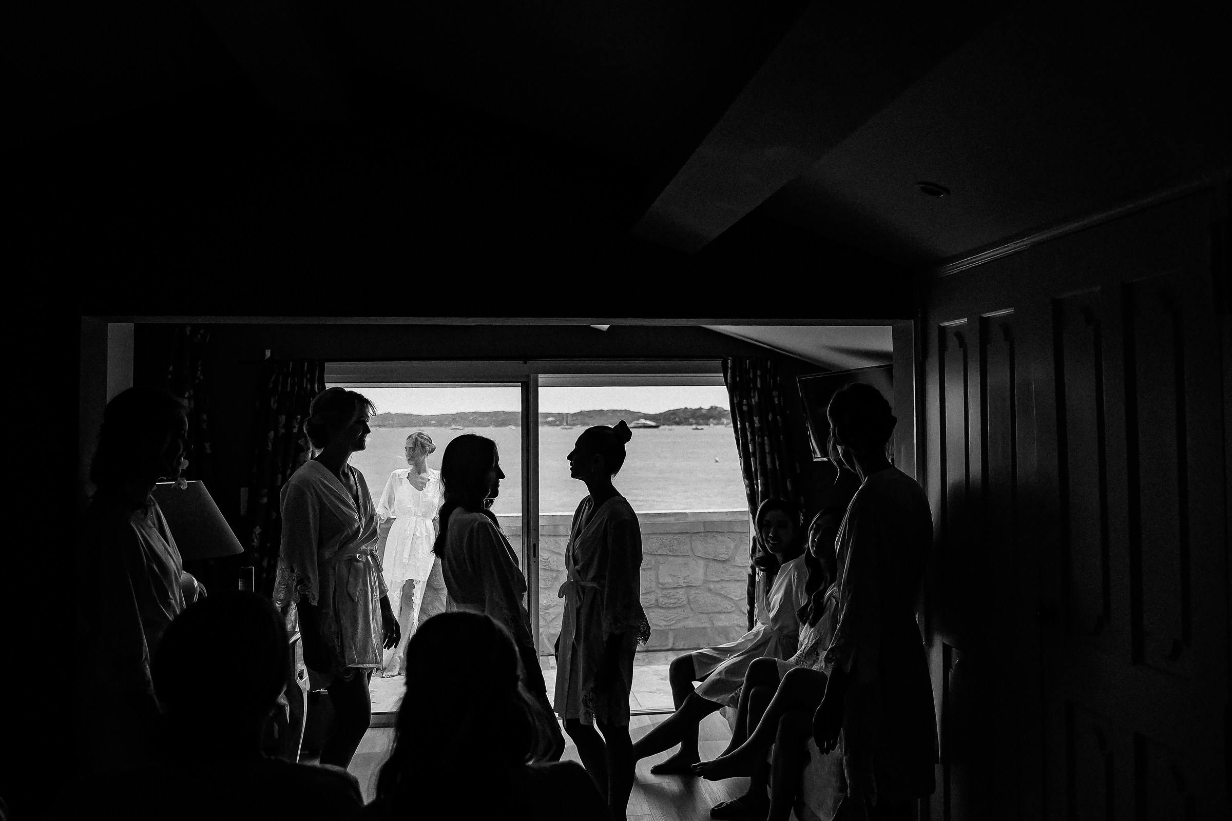 A dramatic silhouette of bridesmaids gathered in a room, overlooking the sea through a wide-open window in Provence.