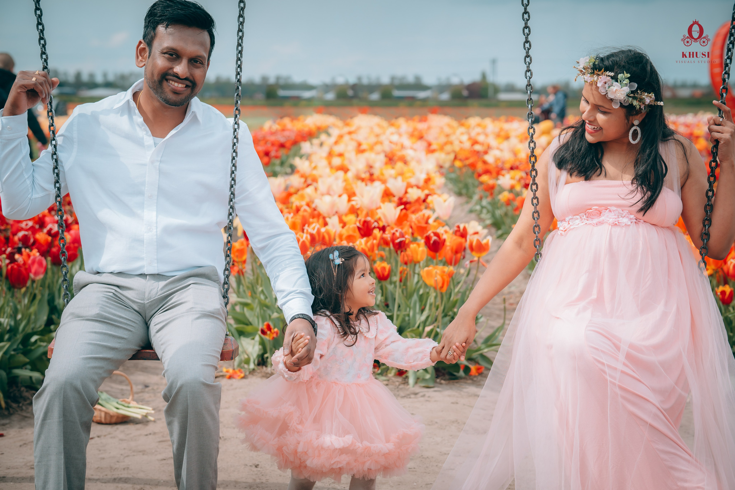 A husband and a pregnant wife sitting on swings and holding hands of their daughter in a tulips fields in netherlands
